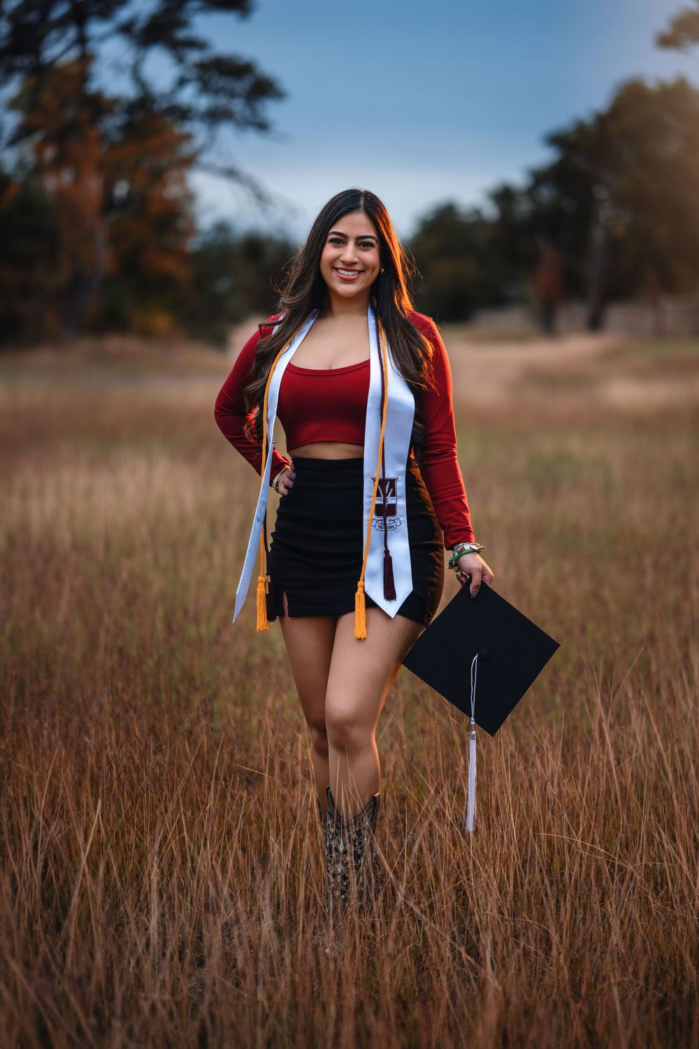 Graduate wearing red crop top and cardigan holds graduation cap while standing in autumn field at sunset.