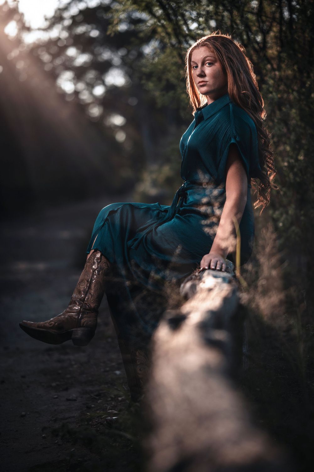 A figure in a teal vintage dress and brown boots sits on a fallen log at sunset, backlit by golden sunlight through trees.