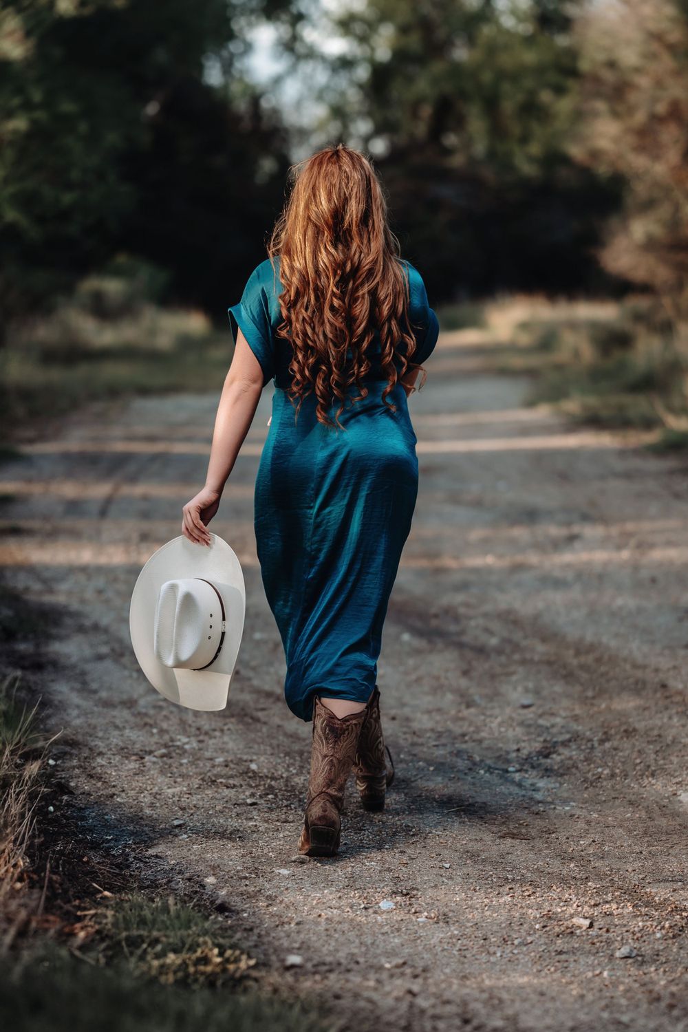 A person in a blue dress and cowboy boots walks down a country path while holding a white western hat.