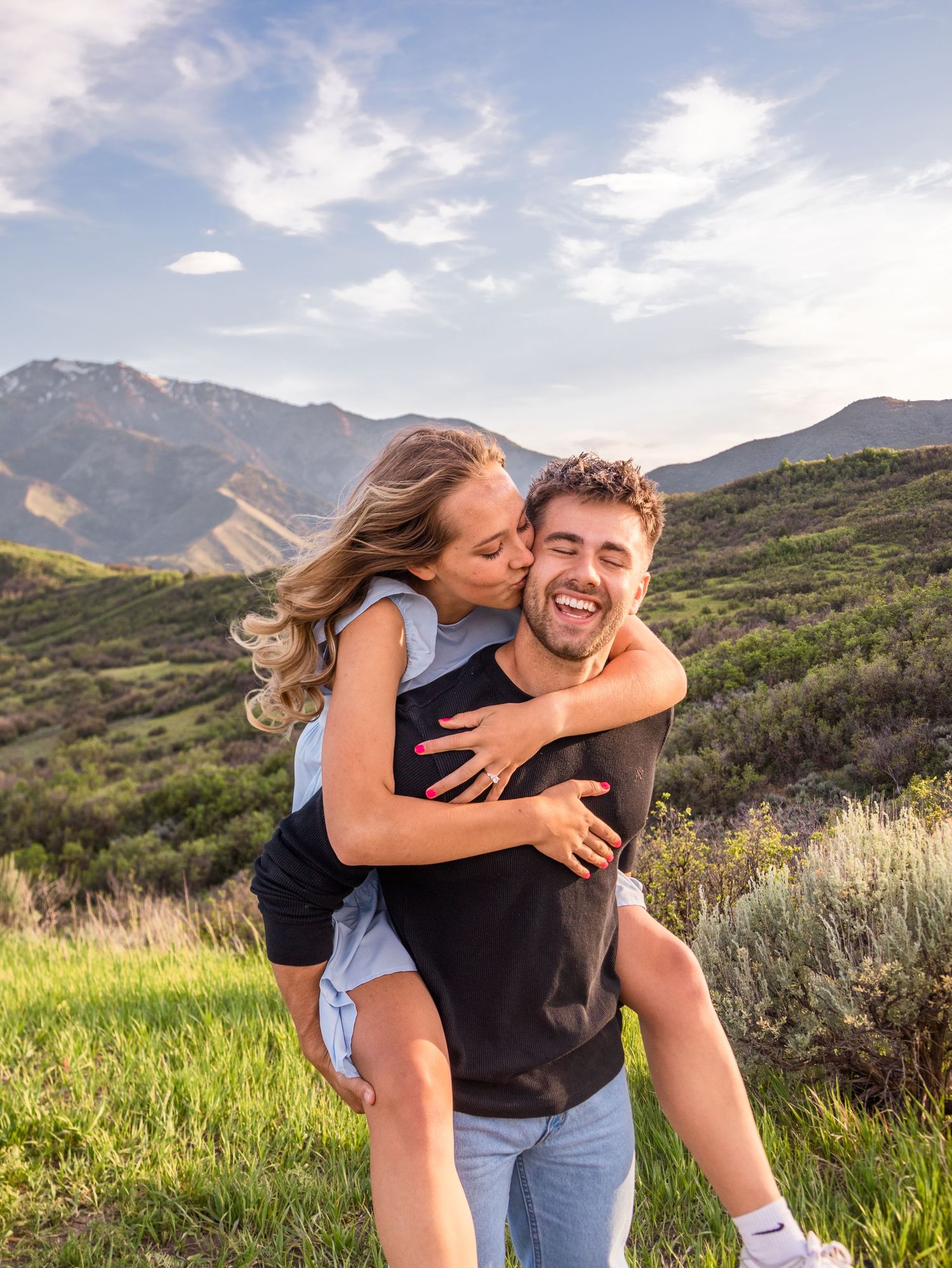 Couple piggyback riding with beautiful Utah green hills behind them for an engagement session