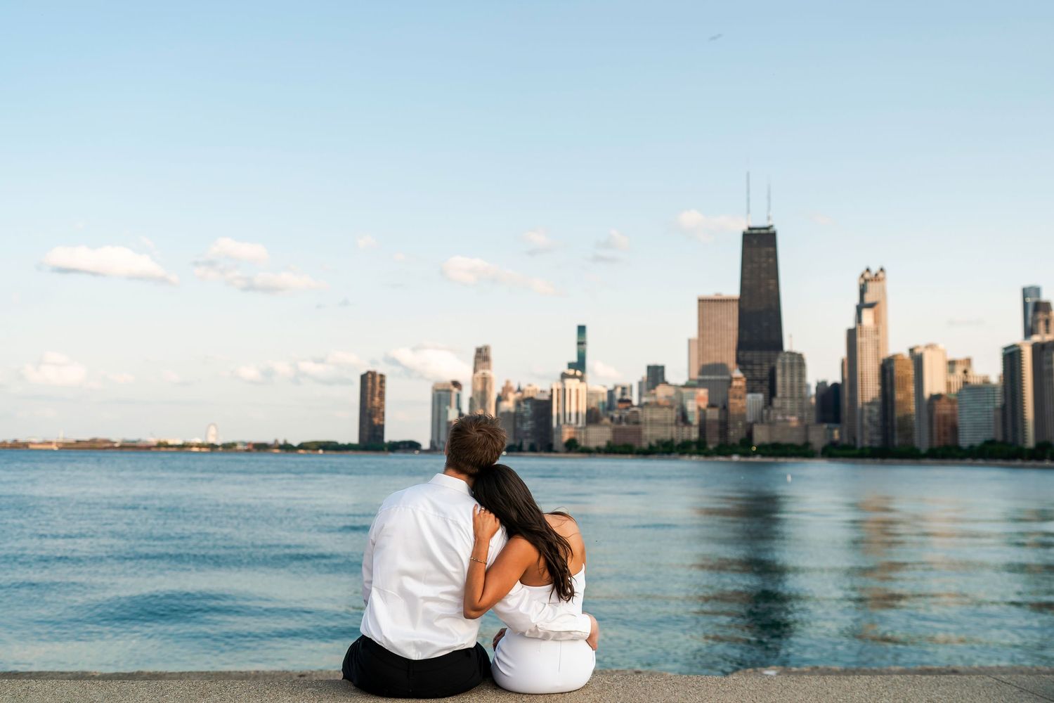 A couple sitting close together on a lakefront ledge, gazing at the Chicago skyline across the water under a soft blue sky.