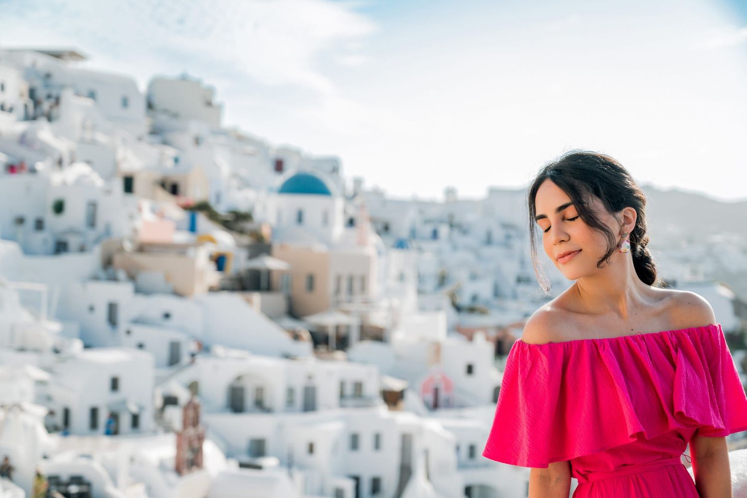 A woman in a pink off-shoulder dress stands against the iconic white buildings and blue domes of Santorini, Greece.