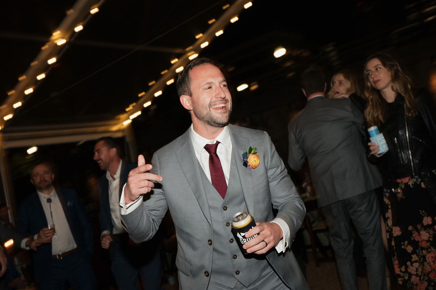 Wedding guest in grey suit and red tie holds a bottle and smiles while mingling at evening reception.