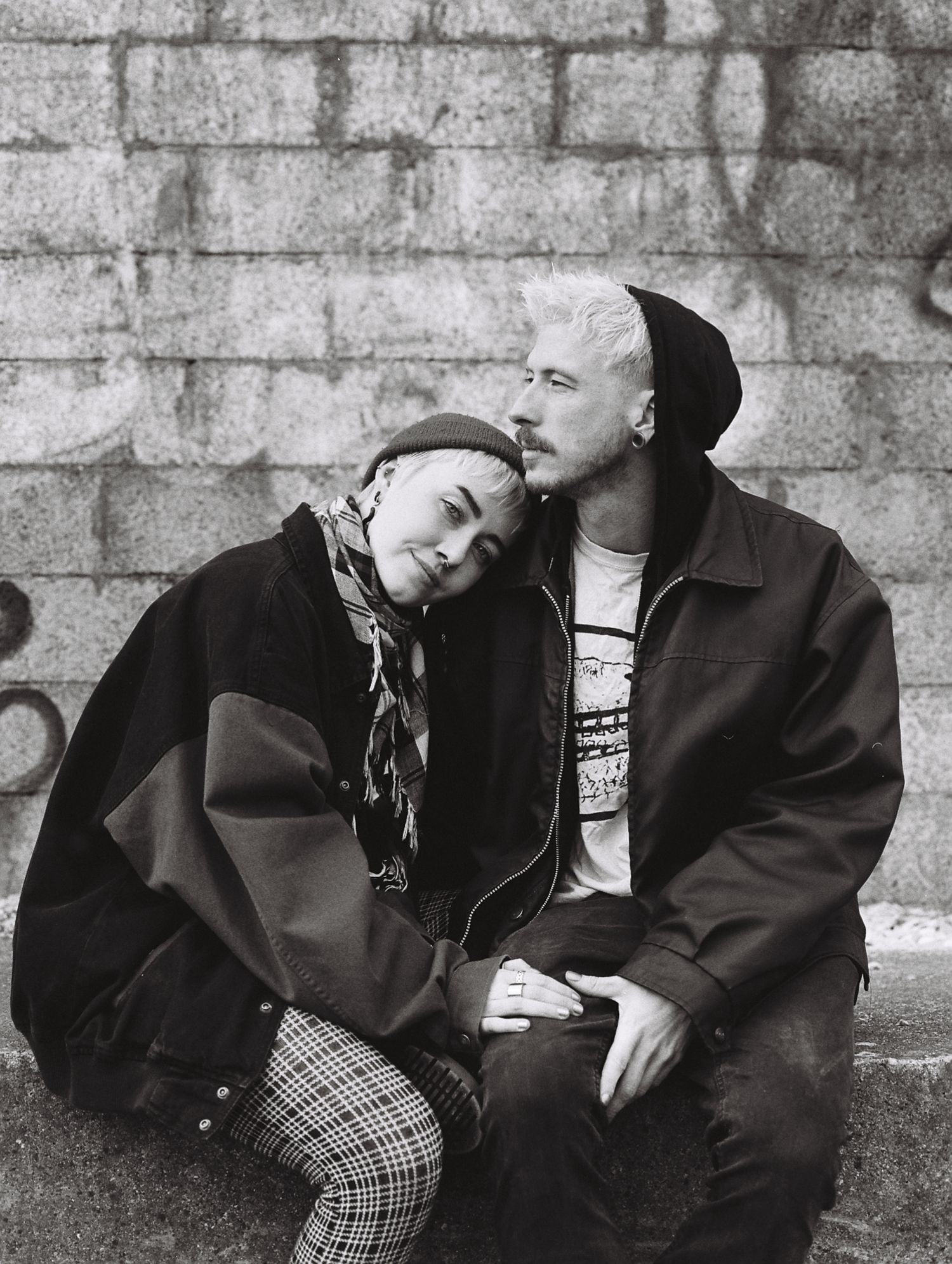 Black and white portrait of a couple sitting against a brick wall wearing casual winter attire.