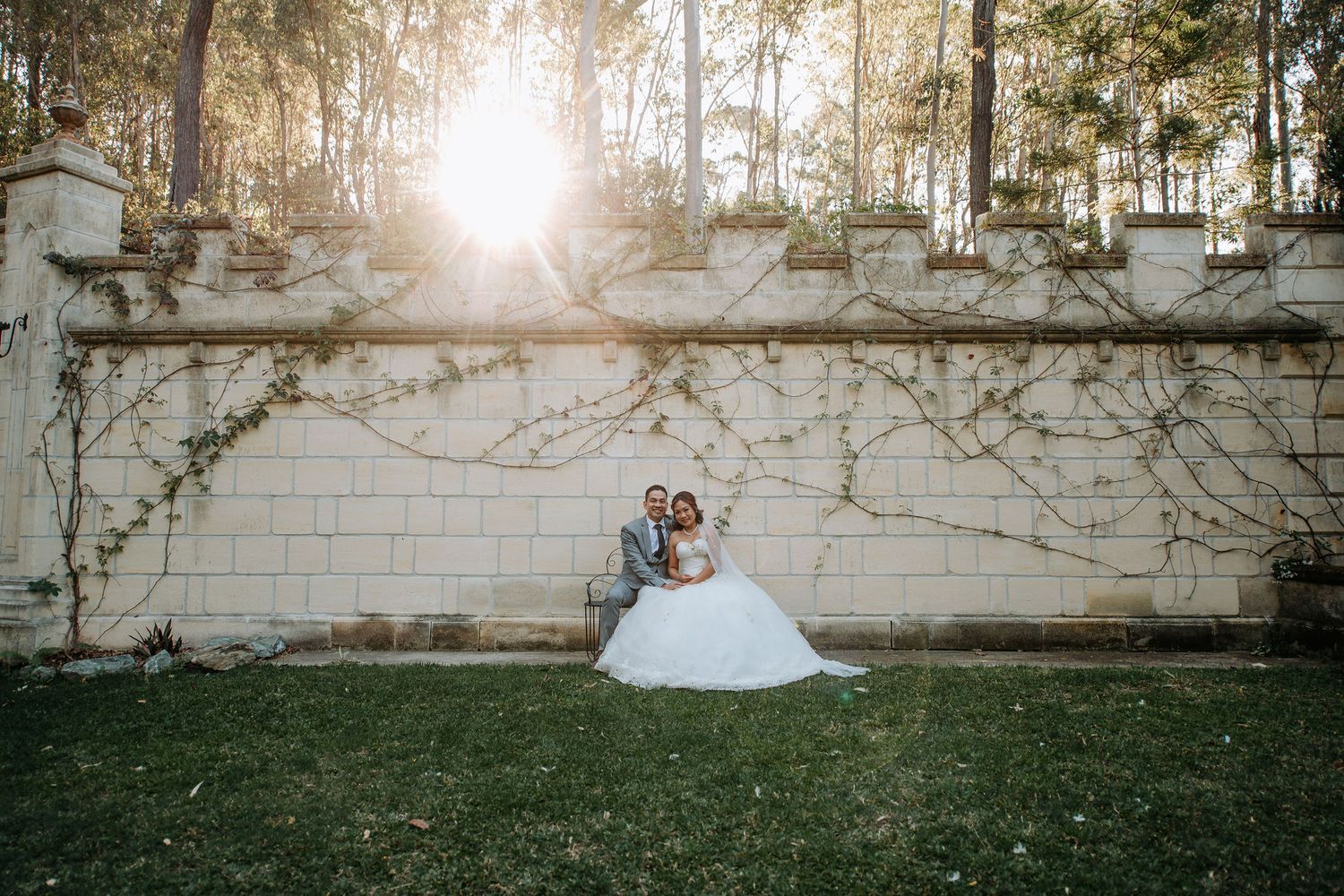 A romantic wedding portrait against a vine-covered wall with sunlight streaming through trees.