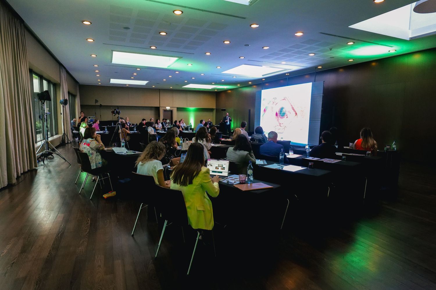 Attendees seated at tables during a professional medical conference presentation in a green-lit hall.