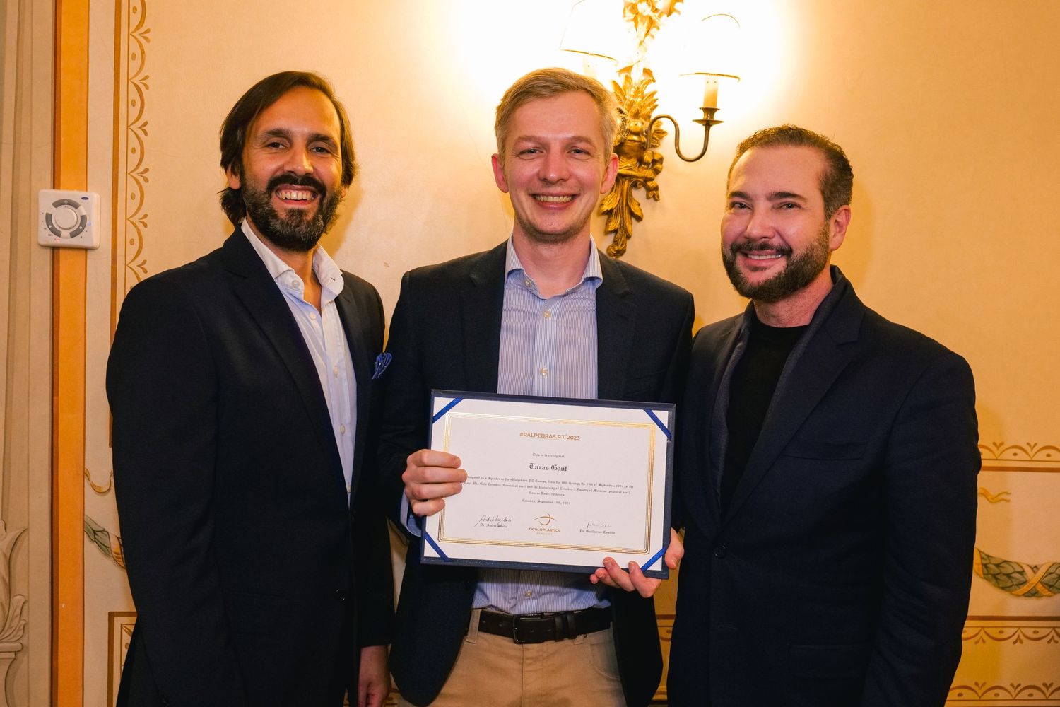 Three men smiling and holding a certificate together at an indoor formal awards ceremony.
