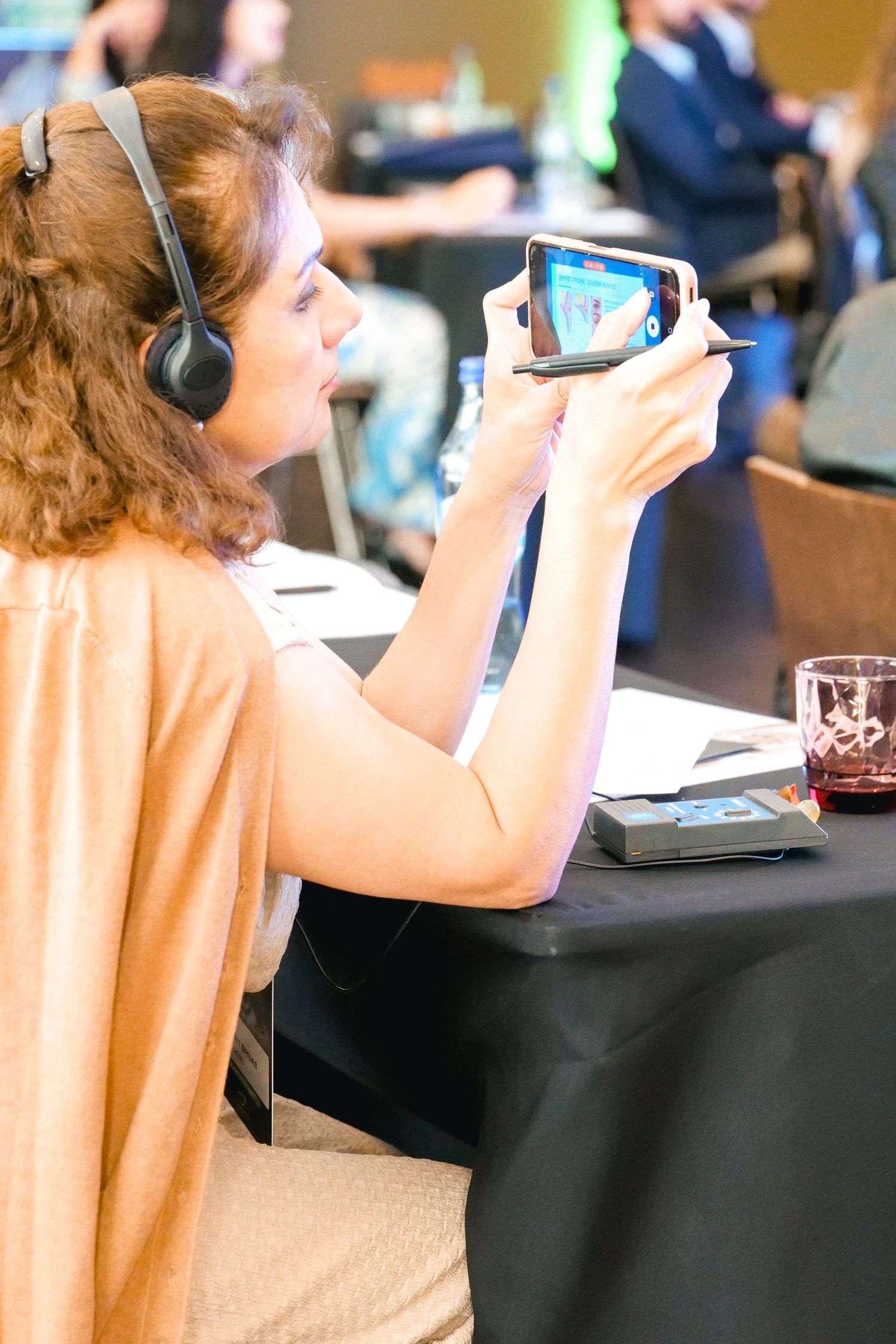 Woman wearing headphones photographing a conference presentation on her smartphone at an event.