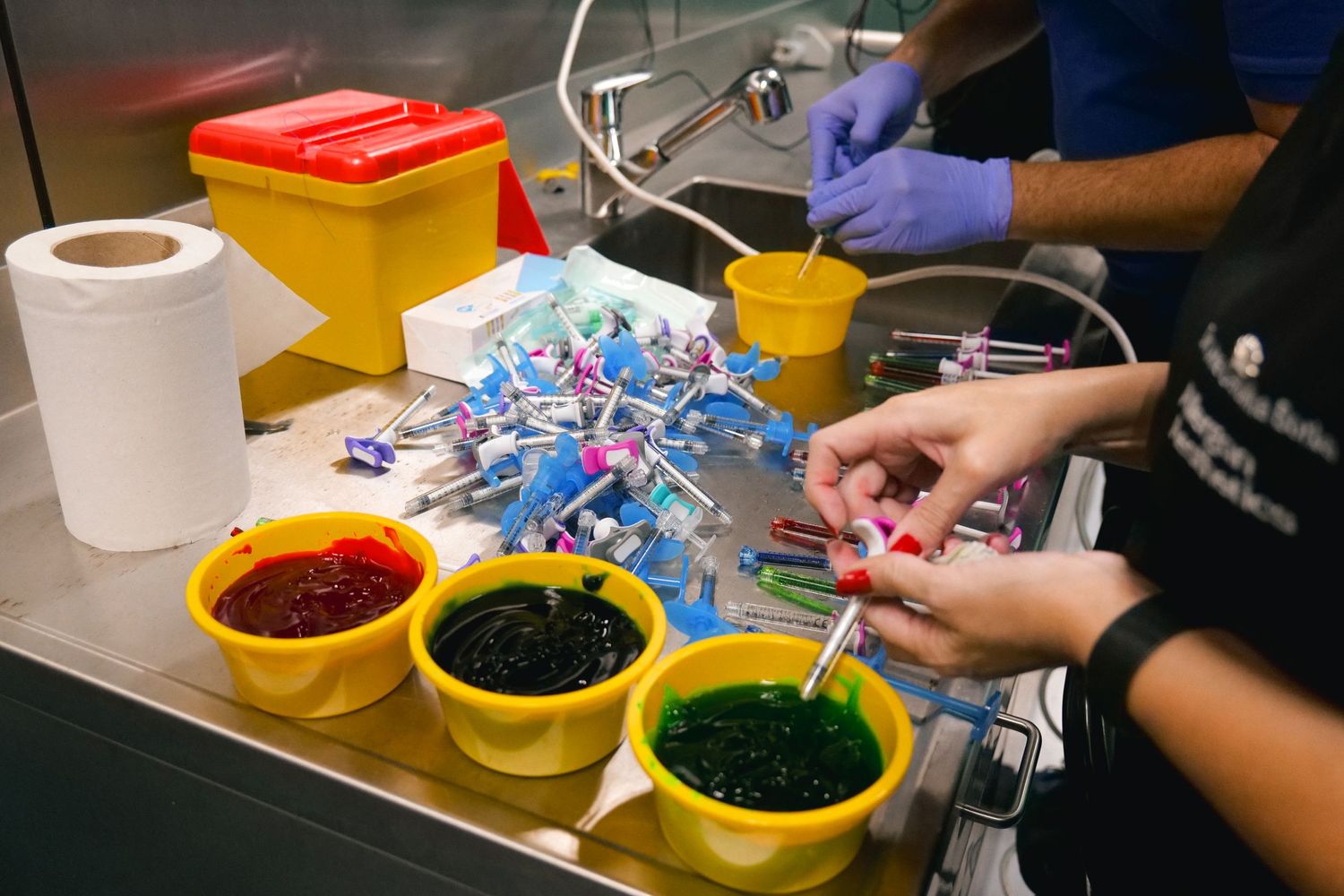 Hands sorting colorful dye-filled syringes into yellow bowls during a medical training exercise.