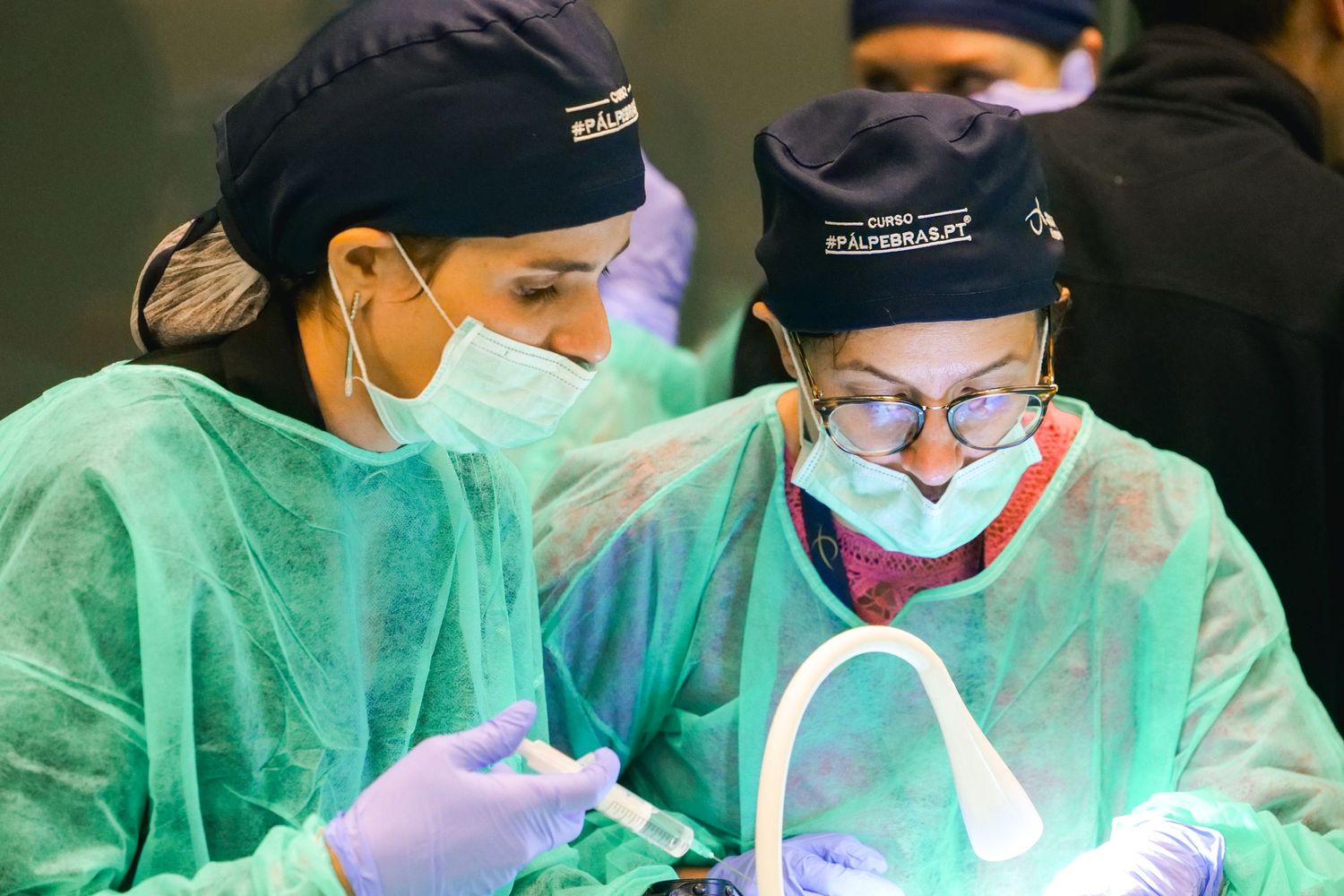 Two female medical professionals in scrubs and masks perform a surgical procedure under bright lighting.