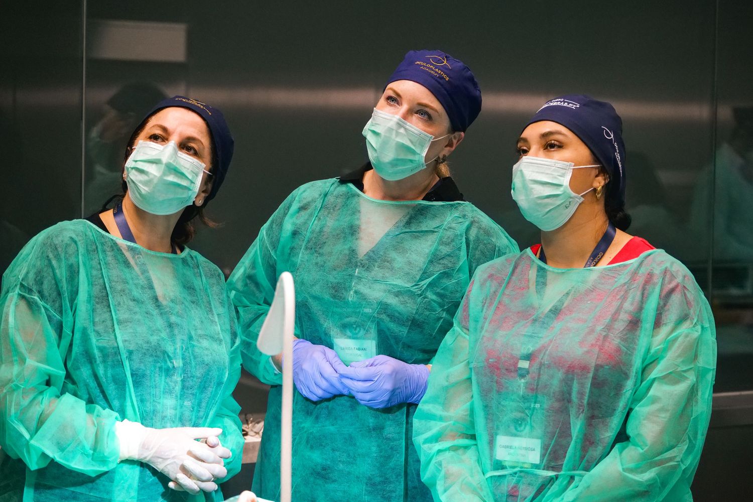 Three female medical professionals in green scrubs and surgical masks stand together in a clinical setting.