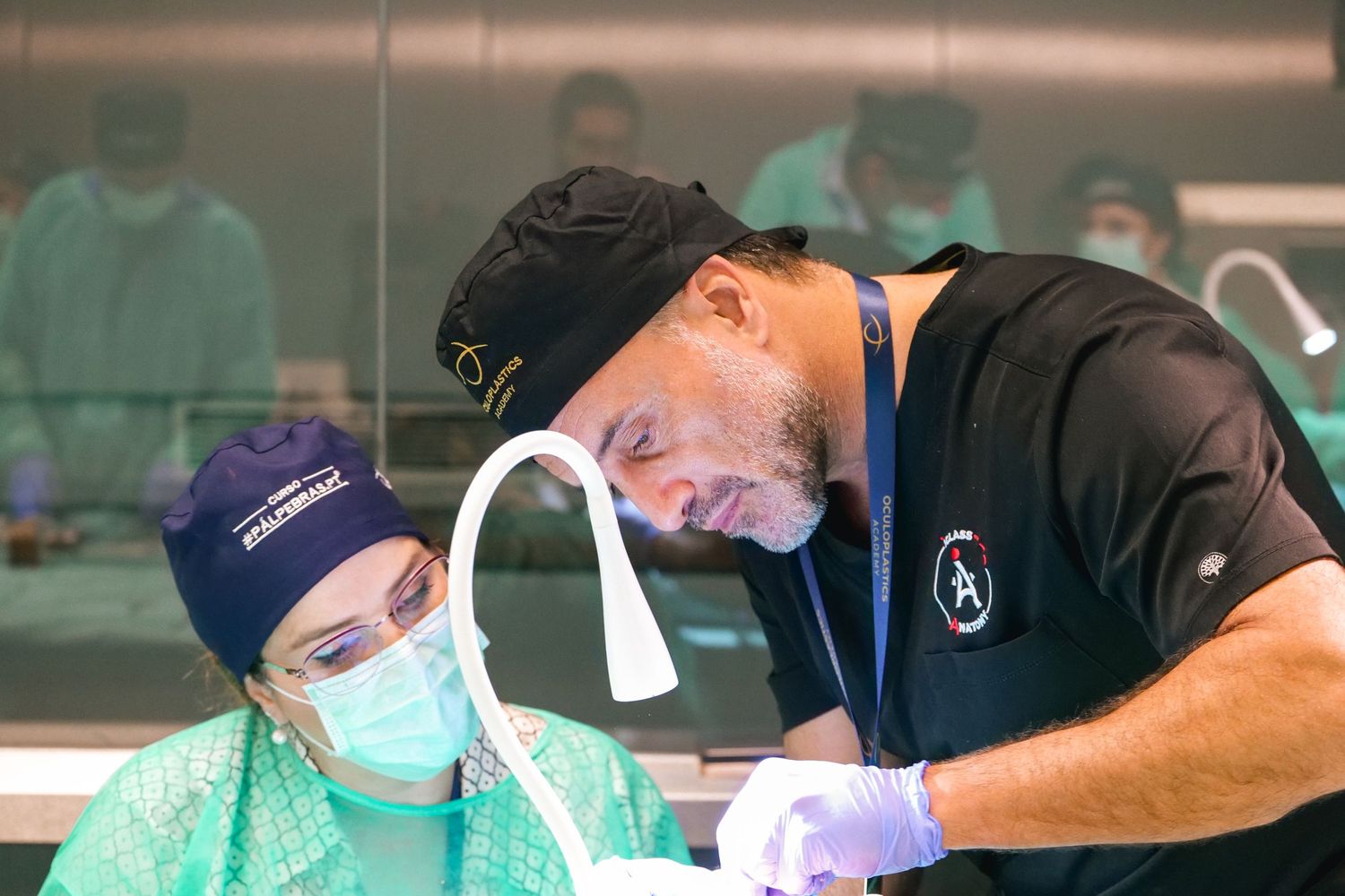 A surgeon in black scrubs guides a masked student during a hands-on medical training procedure.