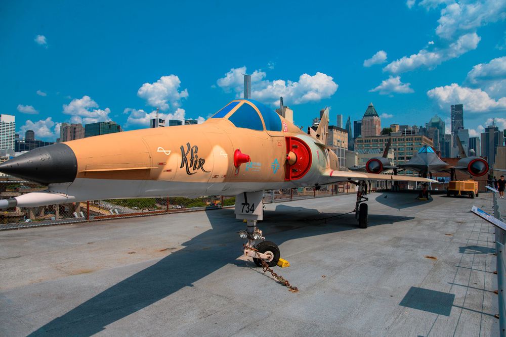 A vintage military aircraft with tan and red paint displayed on a rooftop with a city skyline backdrop.