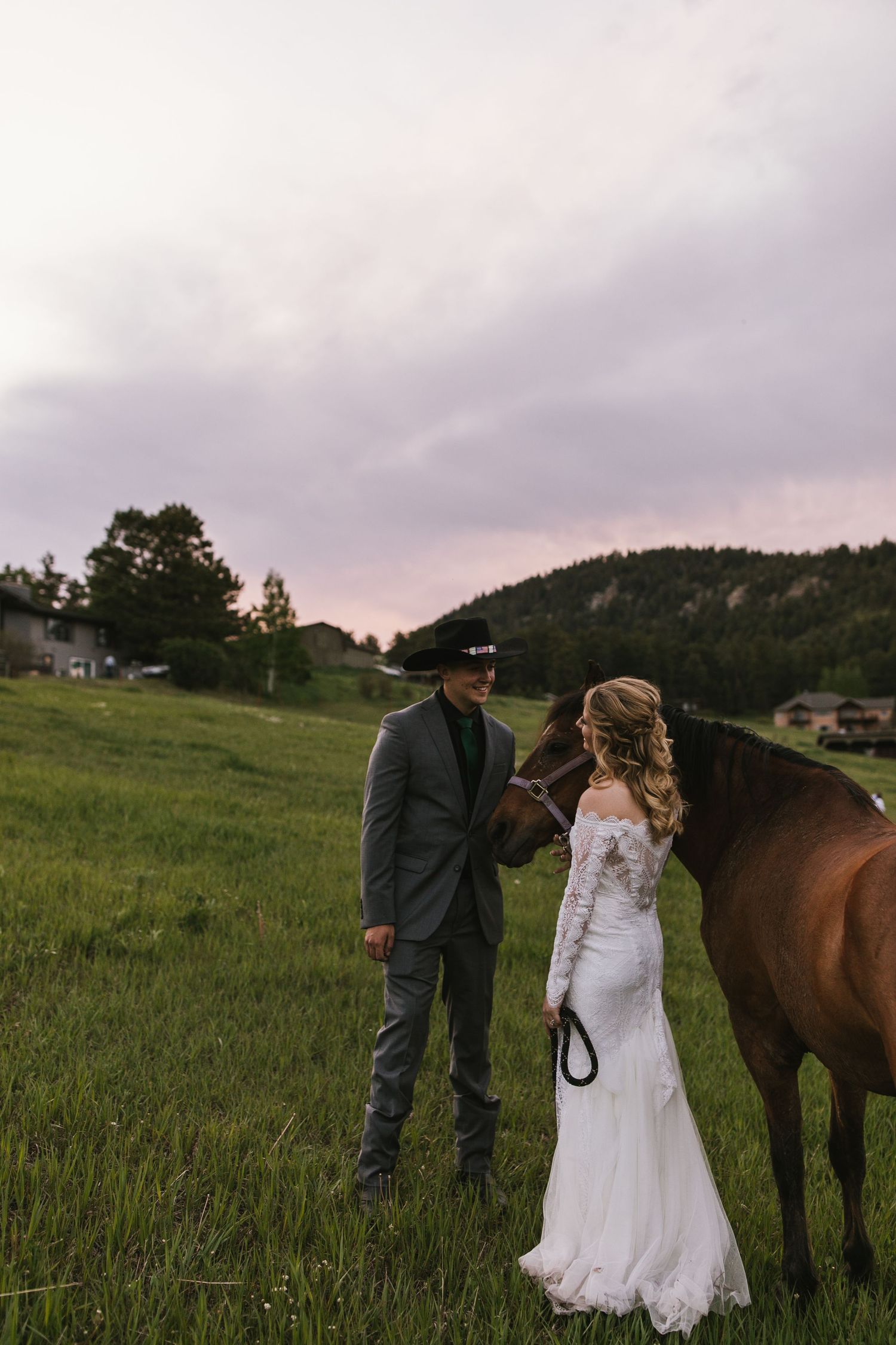 Couple in wedding attire stands with brown horse in rustic mountain setting.