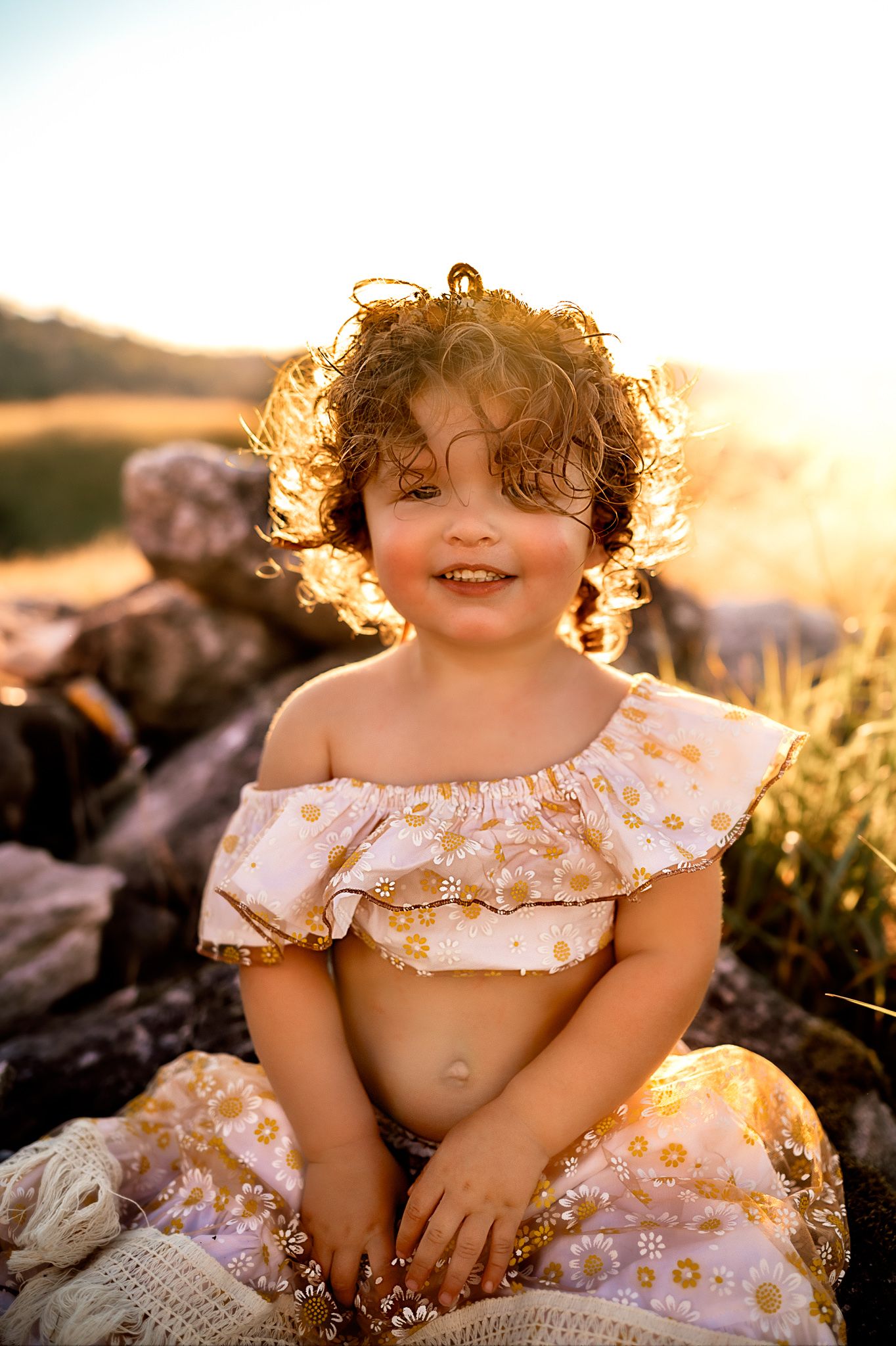 Smiling child with curly hair in floral dress backlit by golden sunset.
