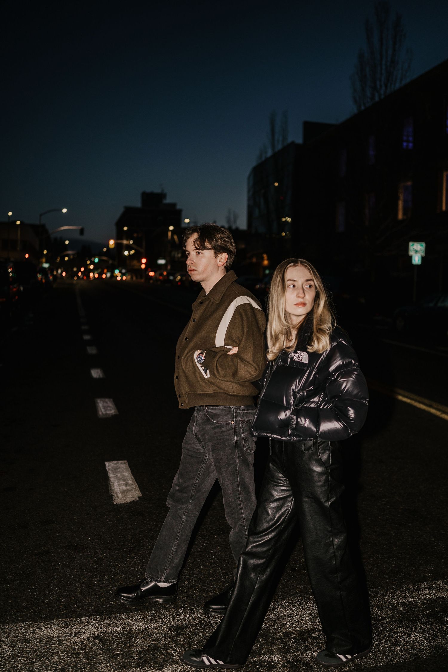 Two people in winter outerwear pose on a dark city street at night with urban lights glowing in the background.