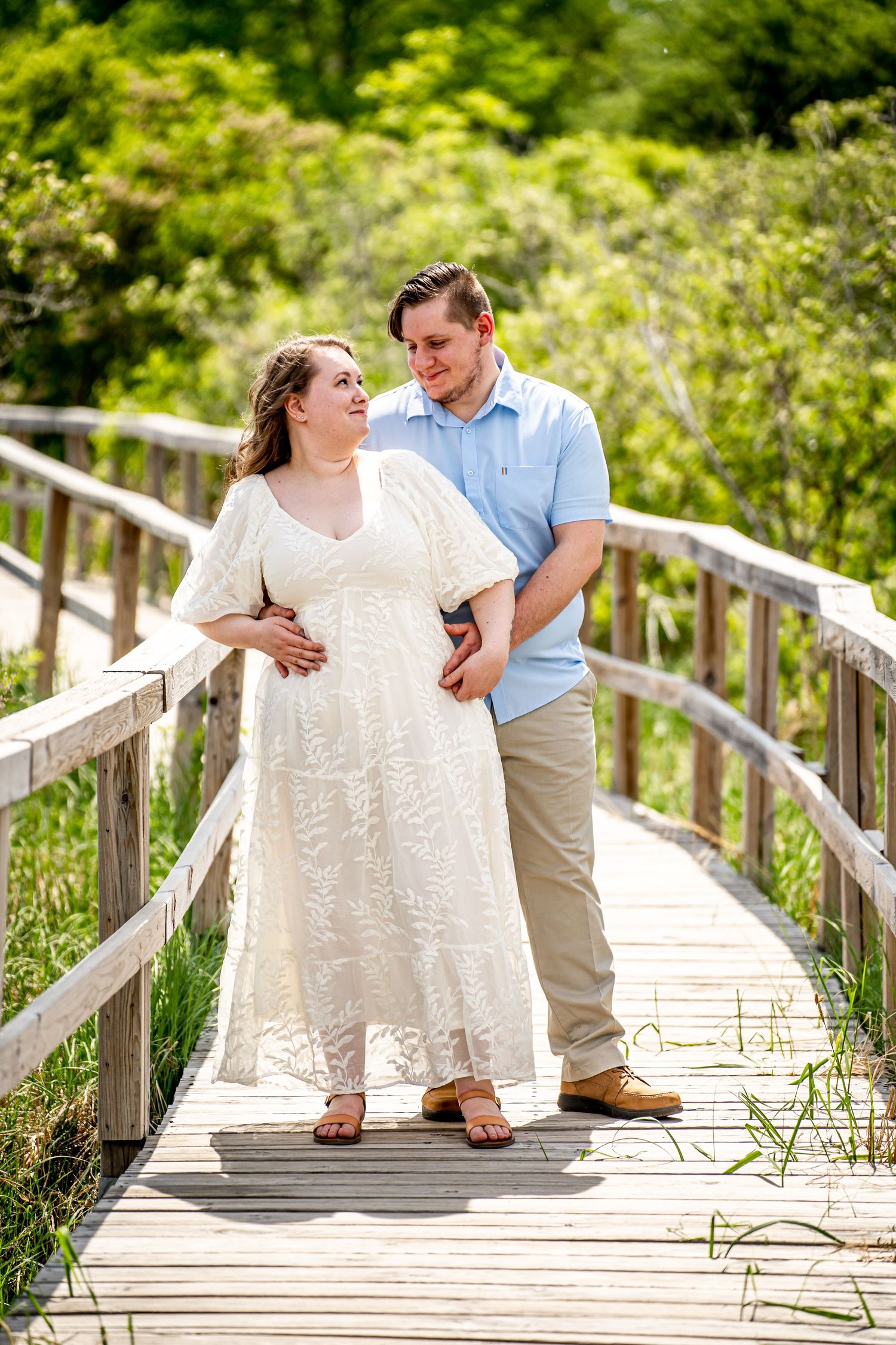 Couple sharing a tender moment on a wooden boardwalk, woman in white lace dress, man in blue shirt, lush green trees.
