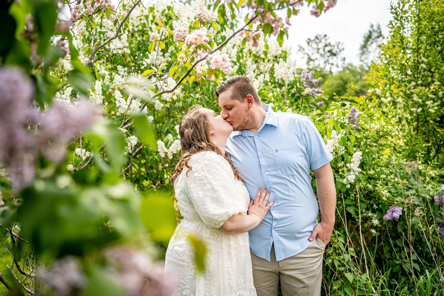 Couple sharing a romantic kiss under blooming spring trees with pink and white flowers in a lush garden setting.