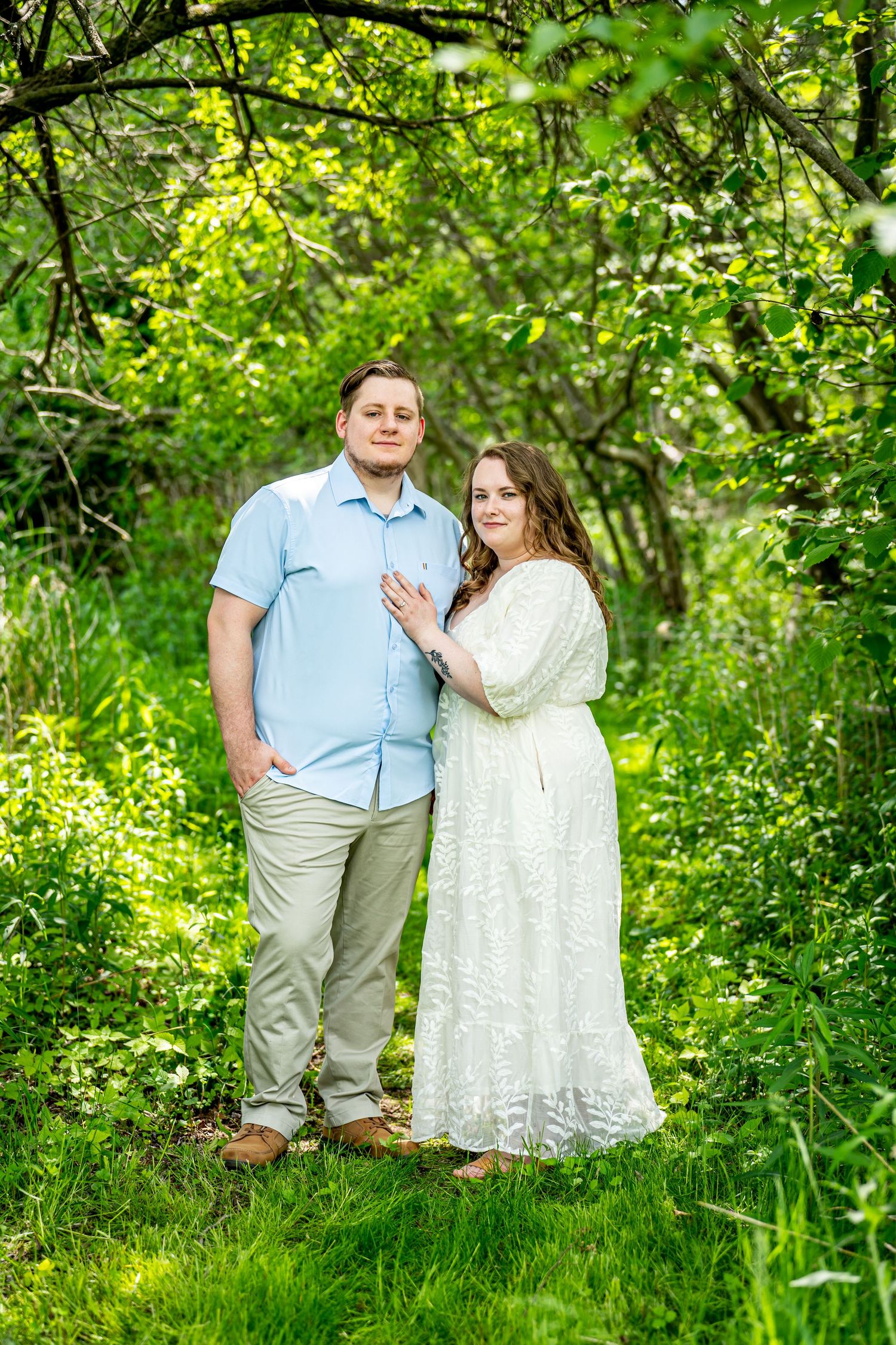 Couple posing outdoors in lush green garden, woman in white lace dress and man in light blue shirt embracing. engagement photos