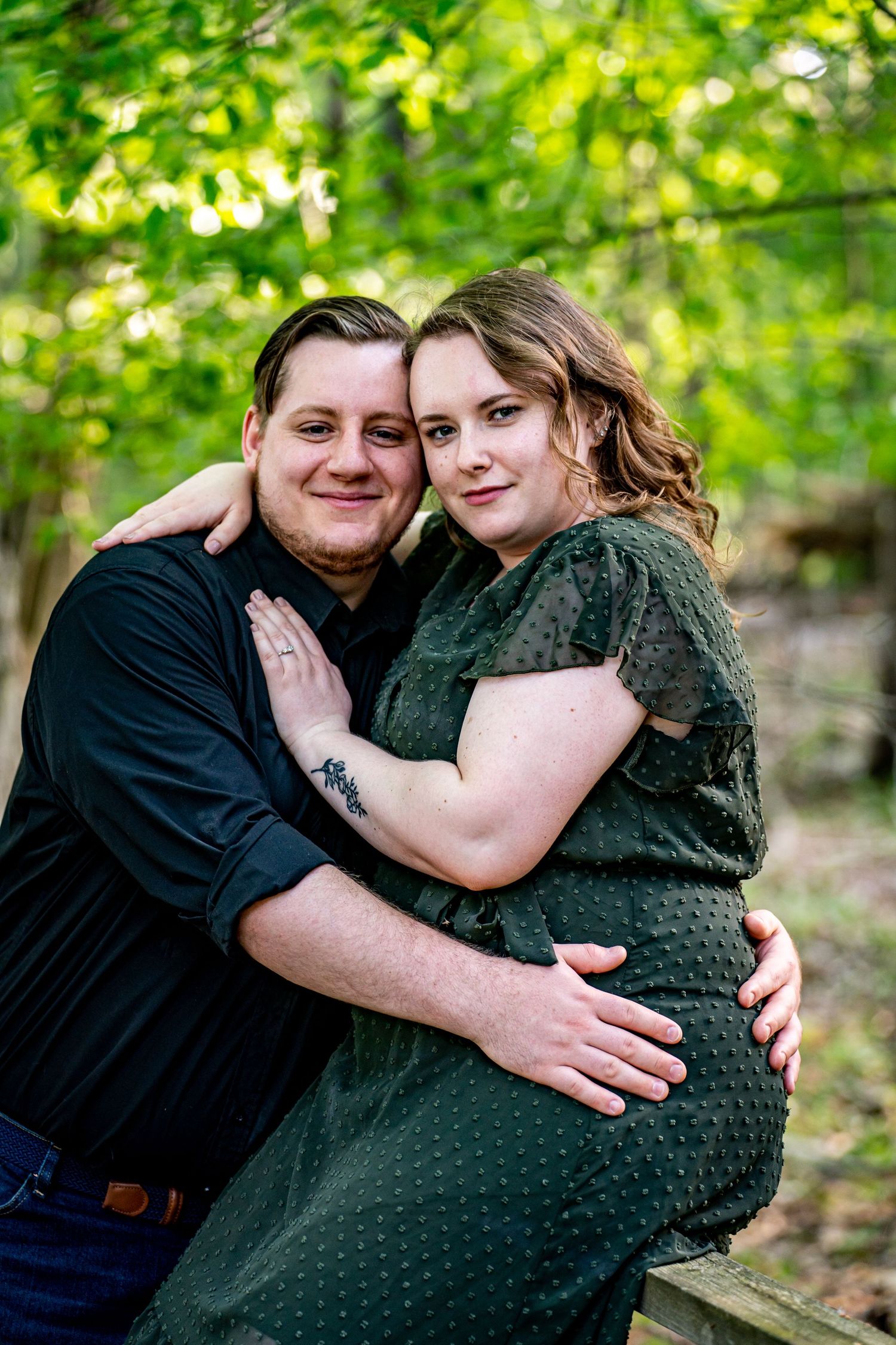 A couple embraces outdoors under green trees, woman in green dress and man in black shirt, smiling warmly. creative couples photography