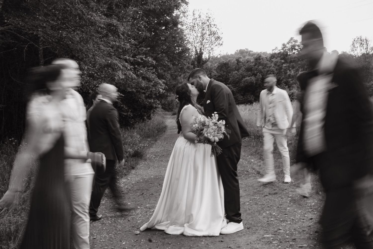 Black and white photo of a wedding couple surrounded by guests on a wooded path.