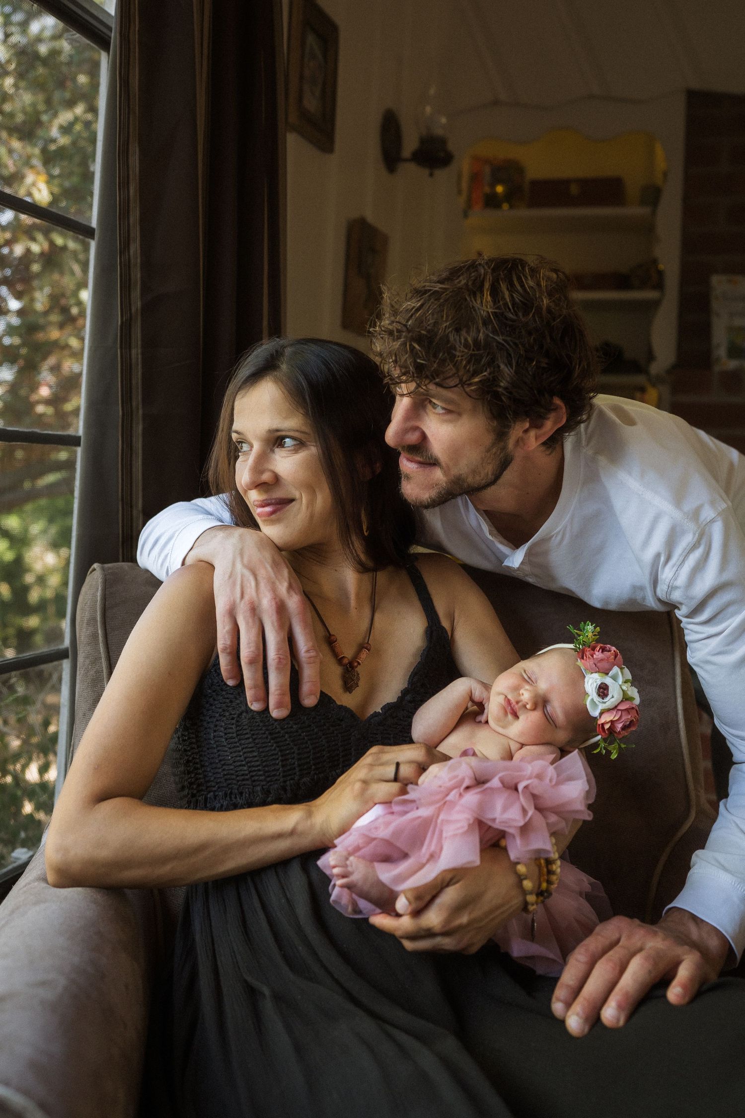 A couple lounges on a couch together while holding their sleeping newborn baby in a pink dress.