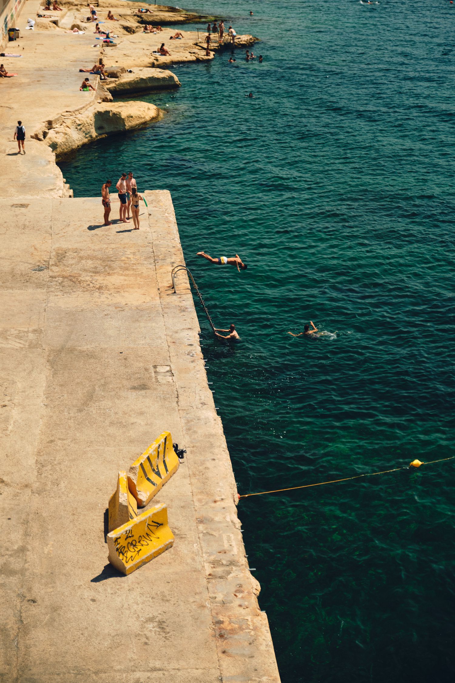 People swimming and relaxing at a concrete pier with turquoise Mediterranean waters.