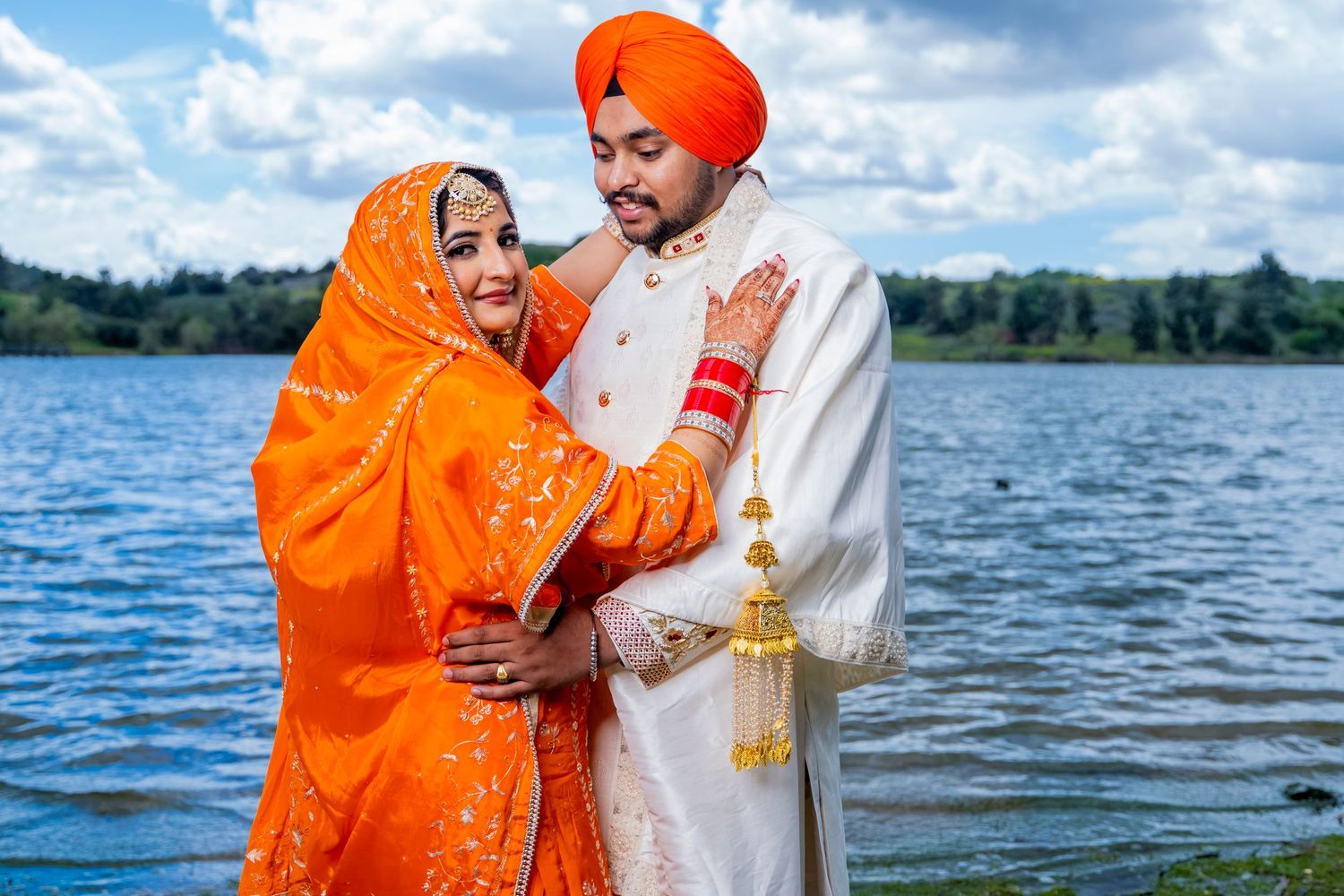 A traditional Sikh couple in orange and white attire pose affectionately by a serene lake. diamond bar.