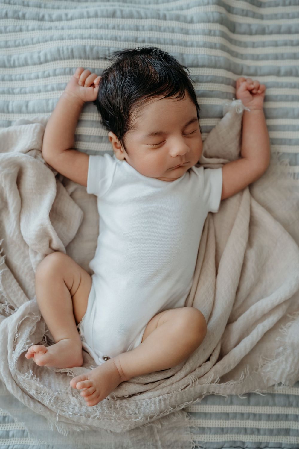 Sleeping baby in white onesie lying on soft textured blankets with arms raised above head.