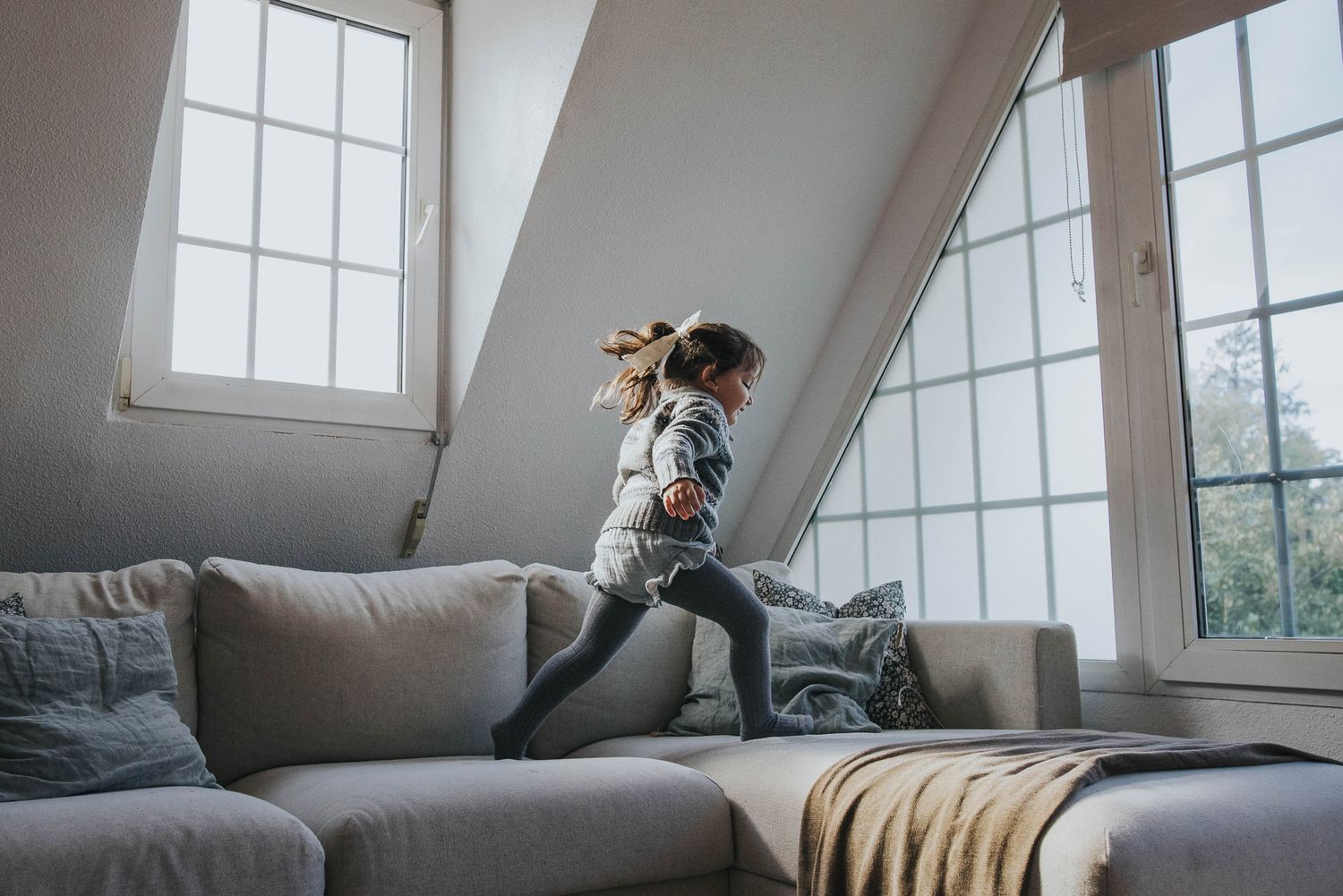 Person lying on gray couch by window in bright attic room while using laptop computer.