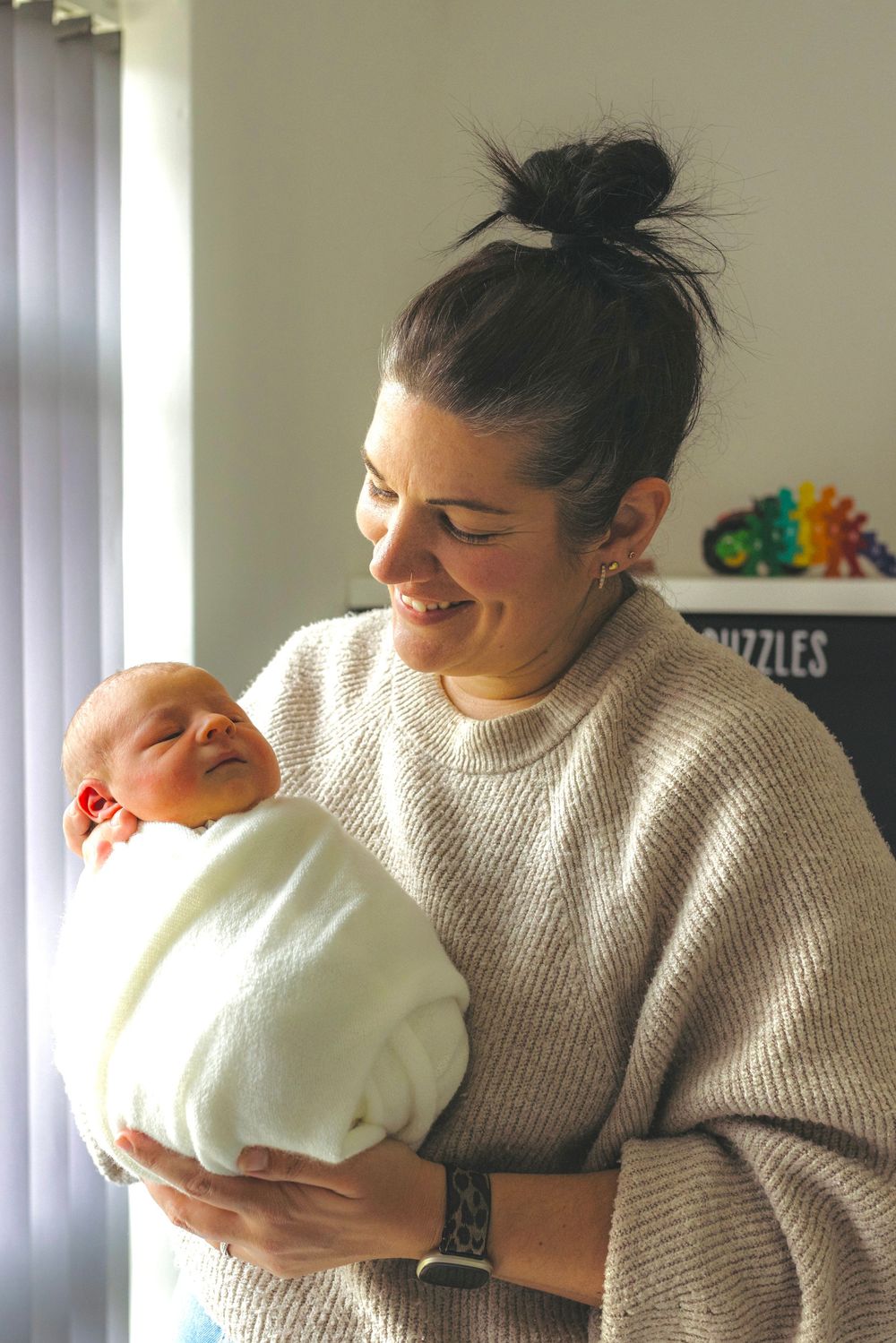 Smiling woman with dark hair in a bun holding a swaddled newborn baby in a white blanket indoors.