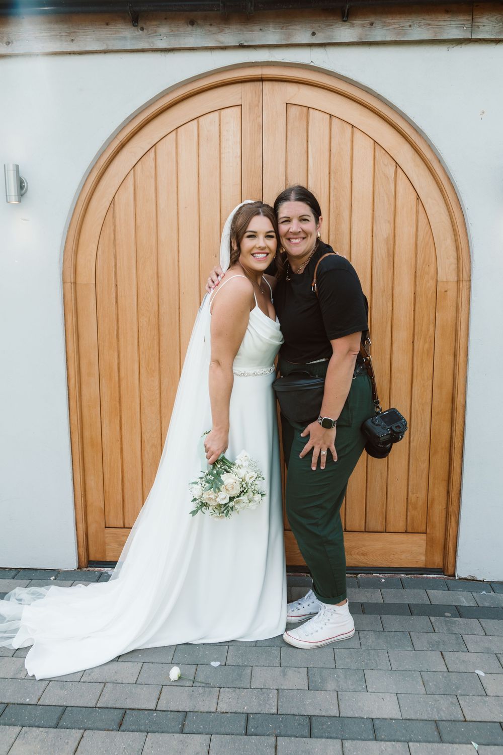 Wedding bride and photographer pose playfully in front of wooden arched doors at outdoor venue. Photographer. Hull. East Yorkshire. Photo