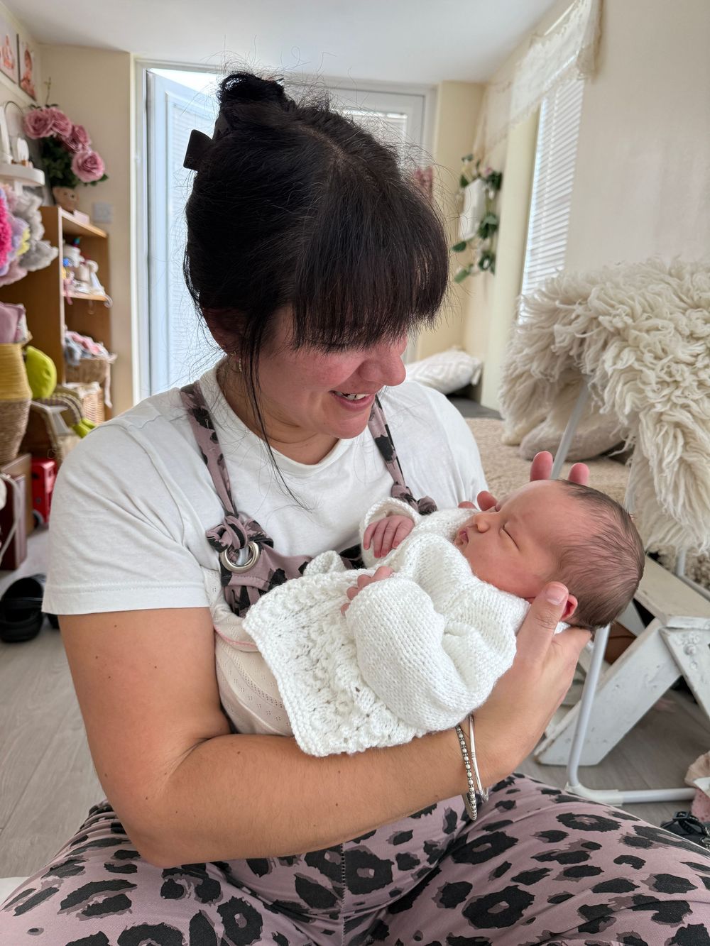 A woman with dark hair smiles down at a newborn baby wrapped in a white knit blanket in a cozy craft room.