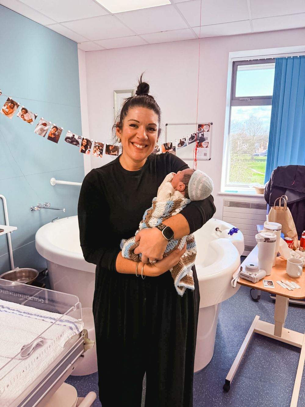 Woman in black outfit smiling while holding a newborn baby in a hospital birthing room.