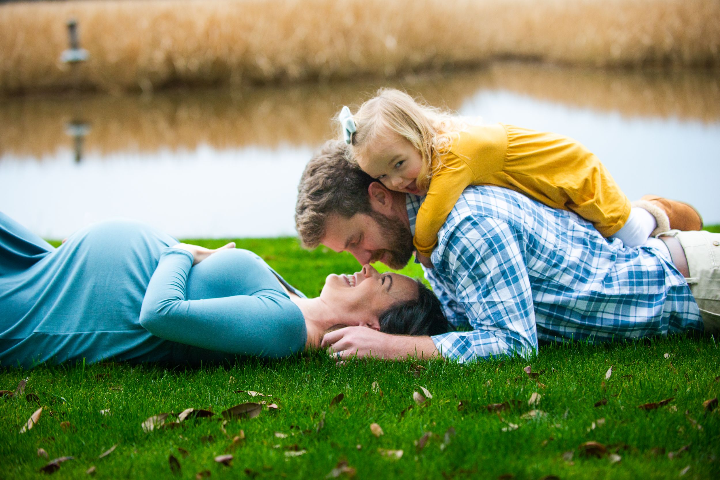 A family lying together on grass by a lake, sharing a tender moment in casual outdoor attire.