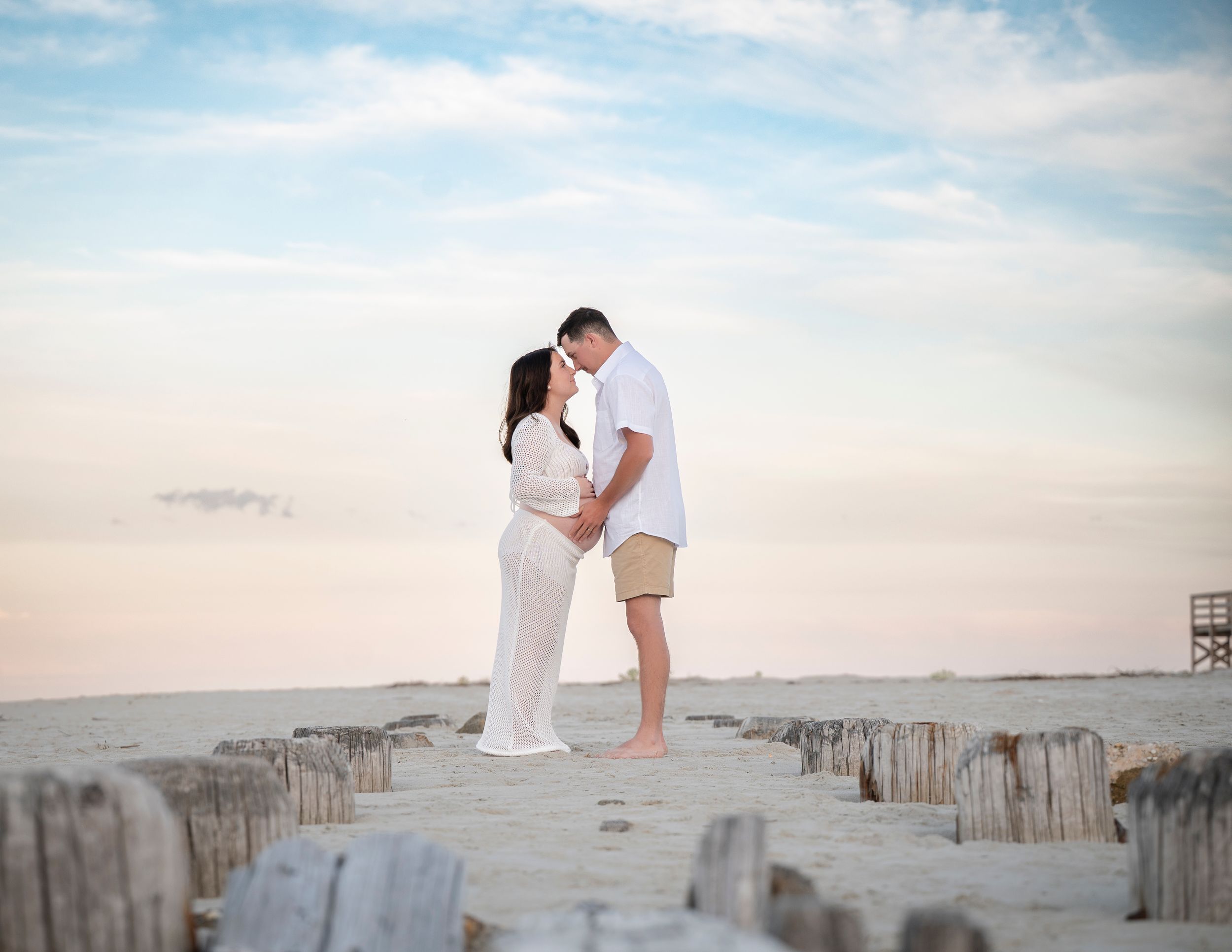 A couple in white embraces on wooden beach posts during sunset, creating a romantic coastal silhouette.