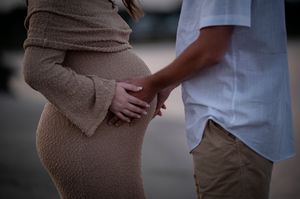 A pregnant person in a beige dress stands close to their partner wearing white as they touch the baby bump tenderly.