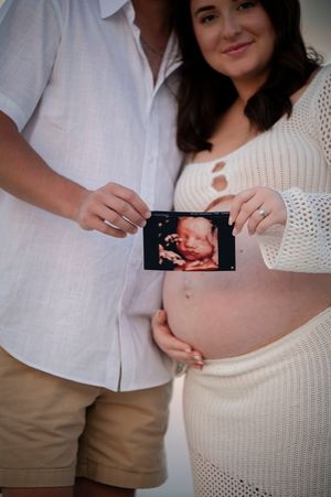 Couple holding sonogram photo against pregnant belly wearing white knit dress and cream colored shirt.