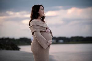 Silhouetted pregnant woman in beige sweater dress stands contemplatively by serene waterfront at dusk.