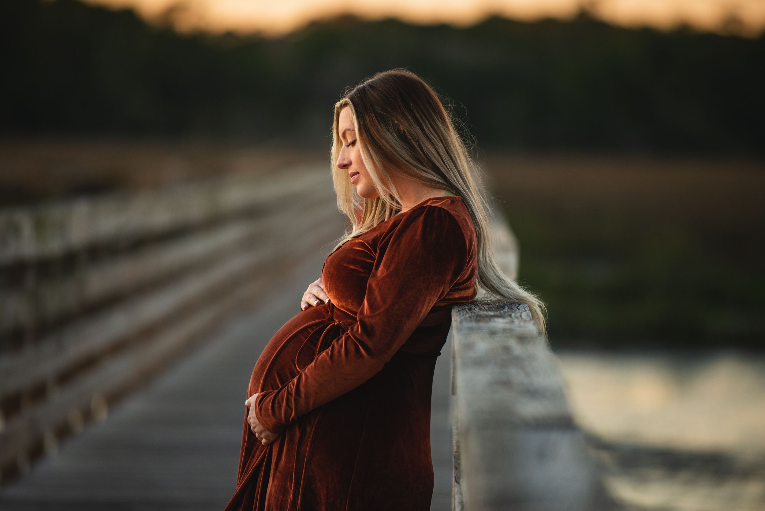 Pregnant woman in maroon dress stands on wooden bridge at sunset.