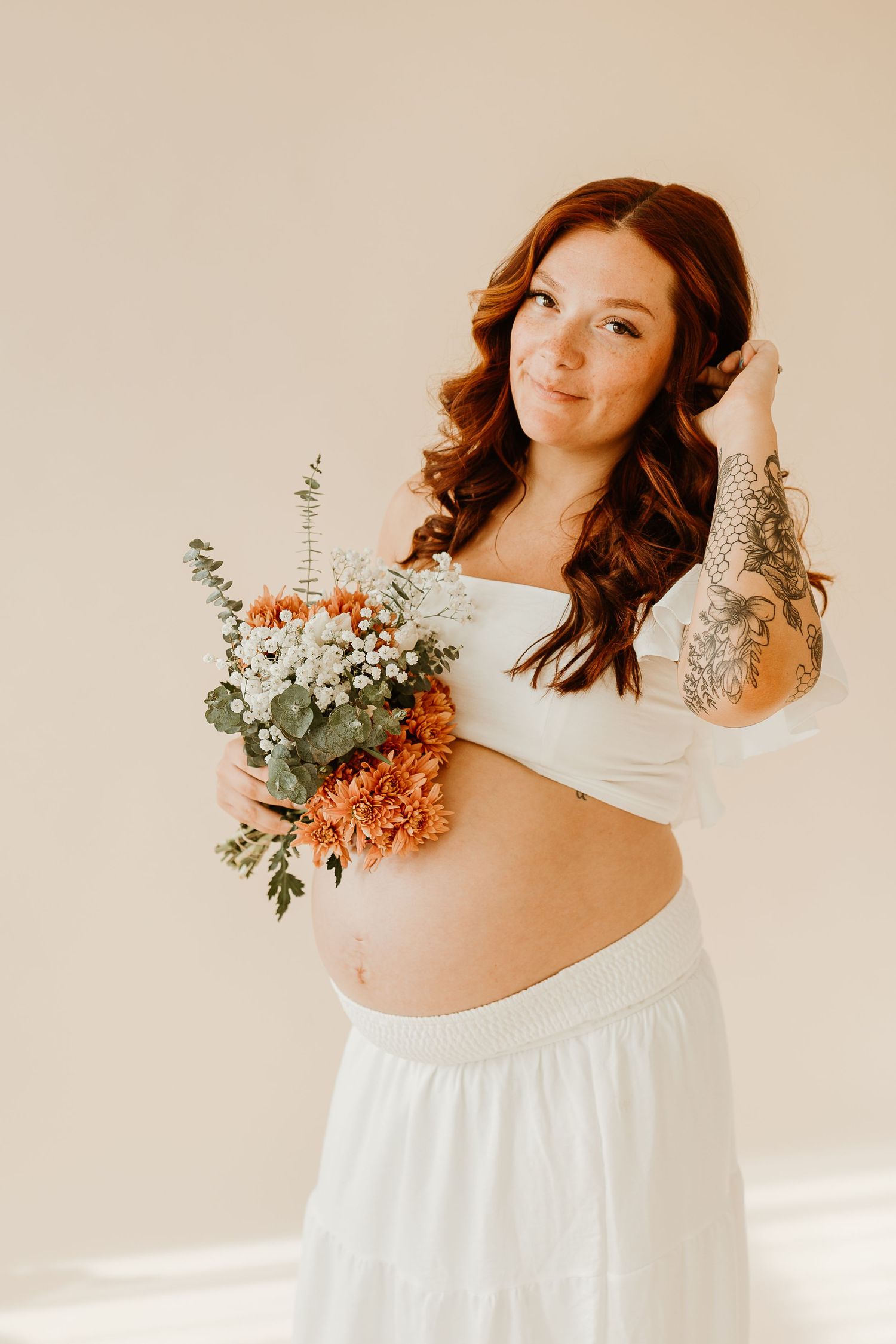 Maternity photoshoot featuring soft white dress and delicate floral arrangements in natural light.