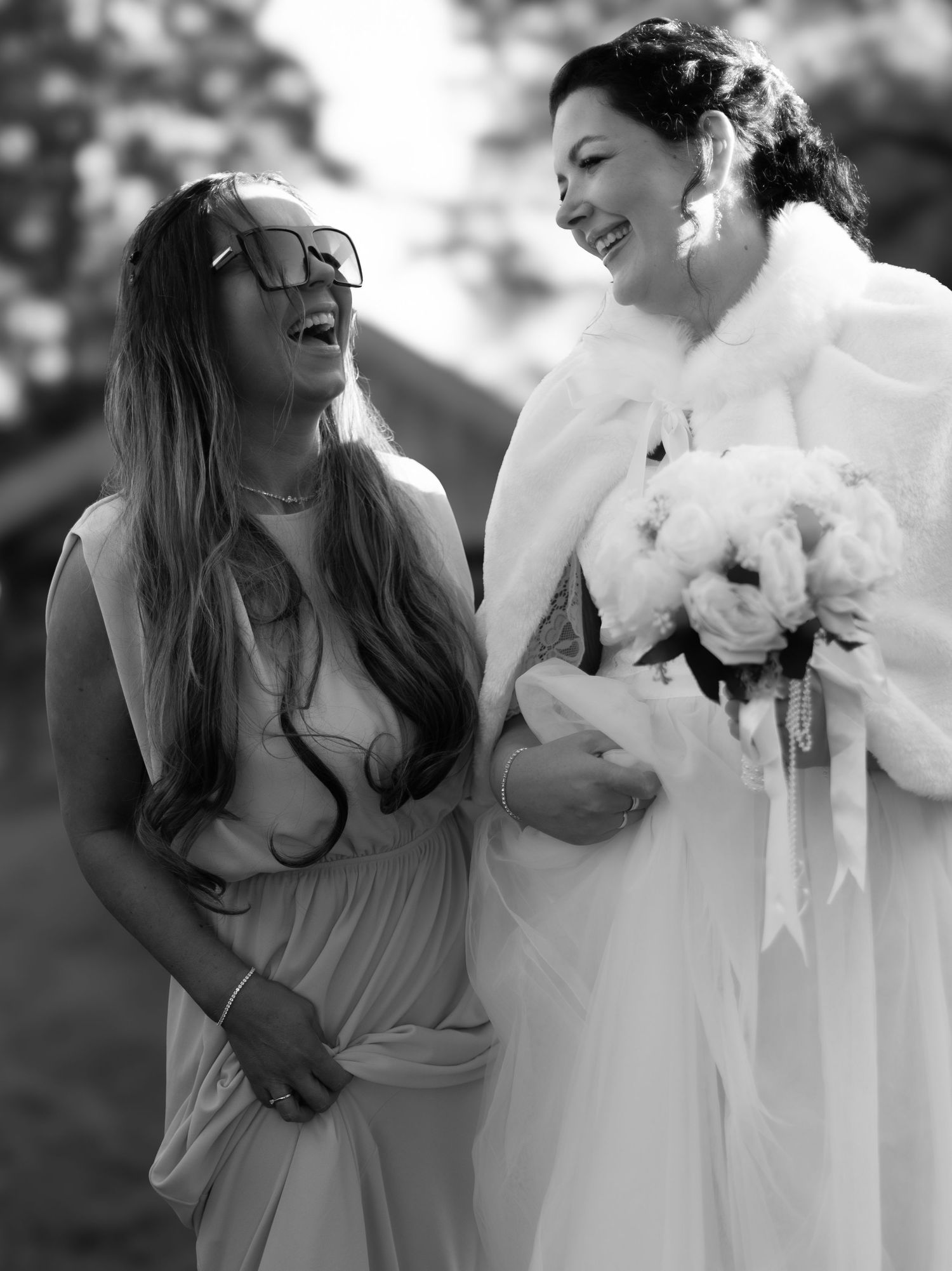 Black and white photo of a bride in white fur shawl and bridesmaid sharing a joyful moment during a wedding.