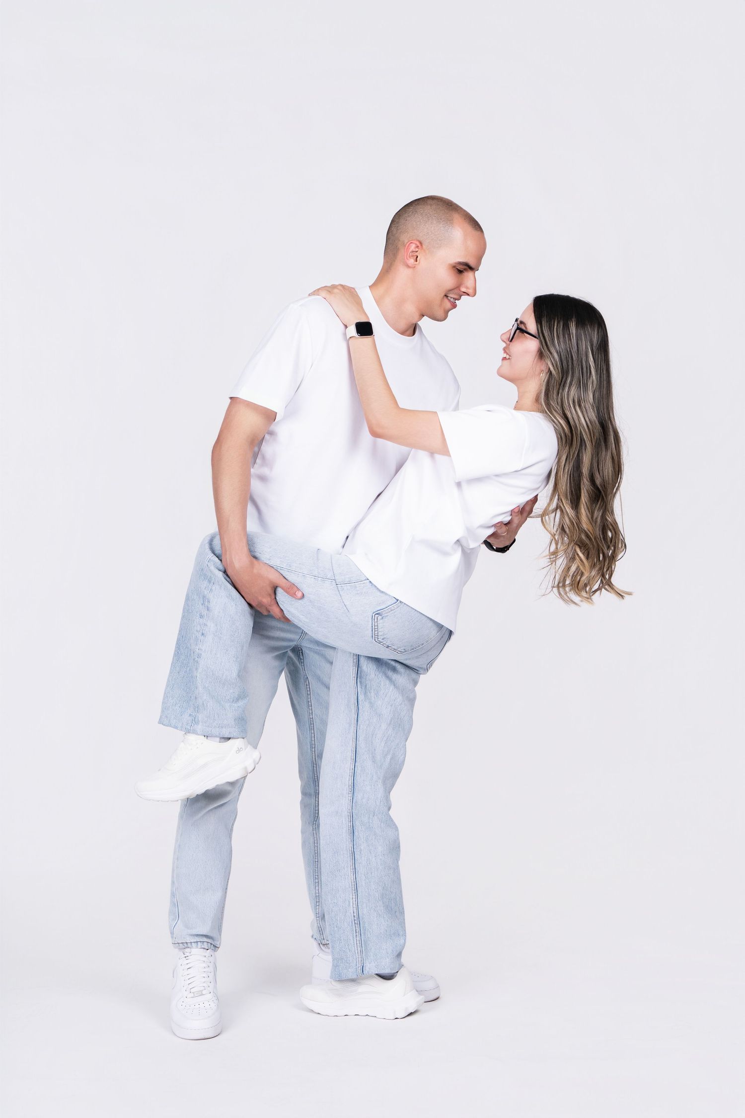 Playful couple in white outfits dancing against white background.