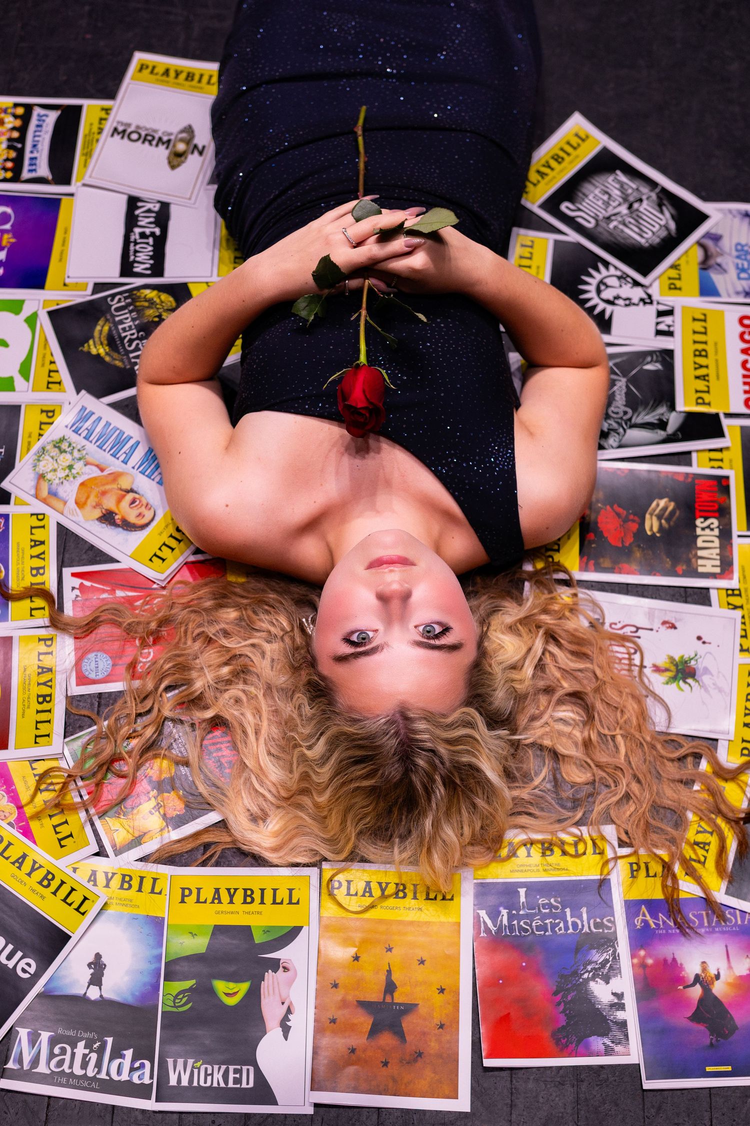 Senior story-telling photographer photo of a girl lying in a bed of playbills holding a rose and looking seriously into the camera.