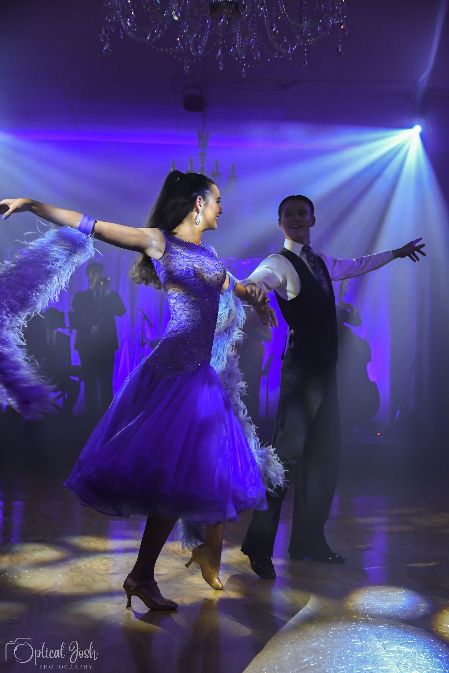 Dancers perform an elegant ballroom dance routine under dramatic purple and blue stage lighting. (Optical Josh)
