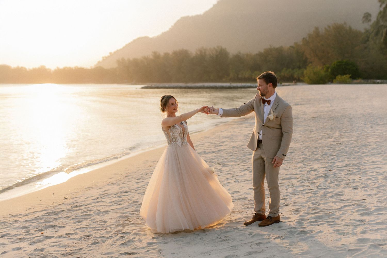 A couple dances together on a sandy beach at sunset with mountains and ocean in the background.