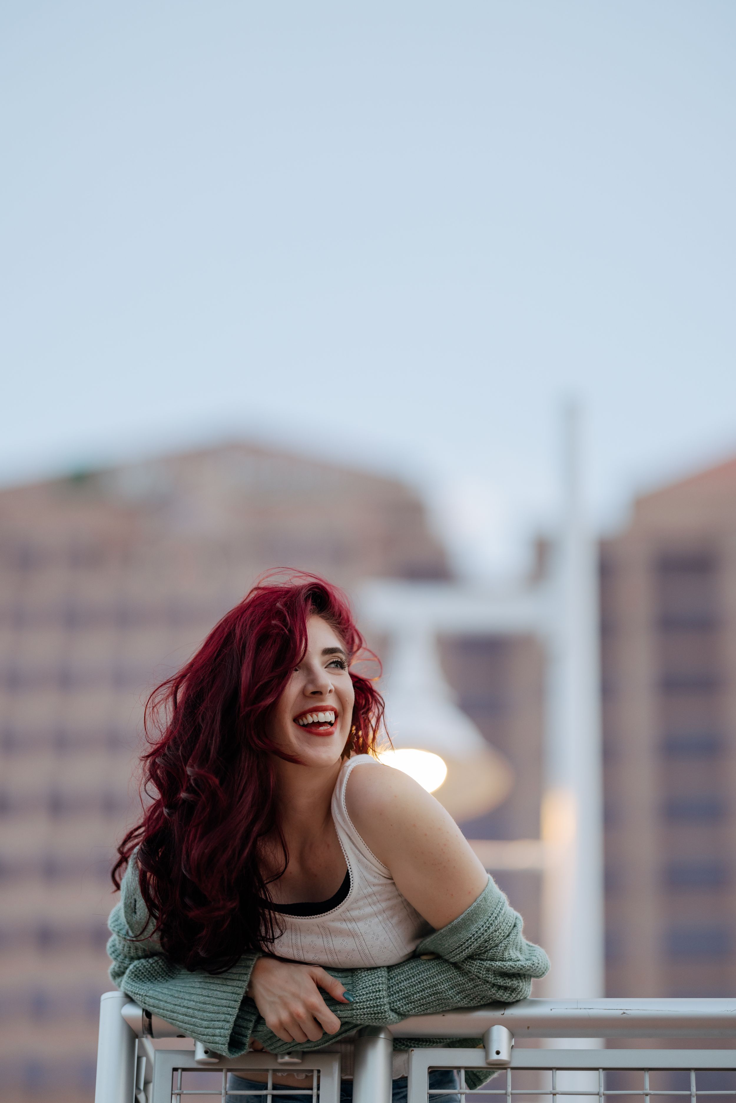 Woman with vibrant red hair laughing while leaning on balcony railing against city backdrop.