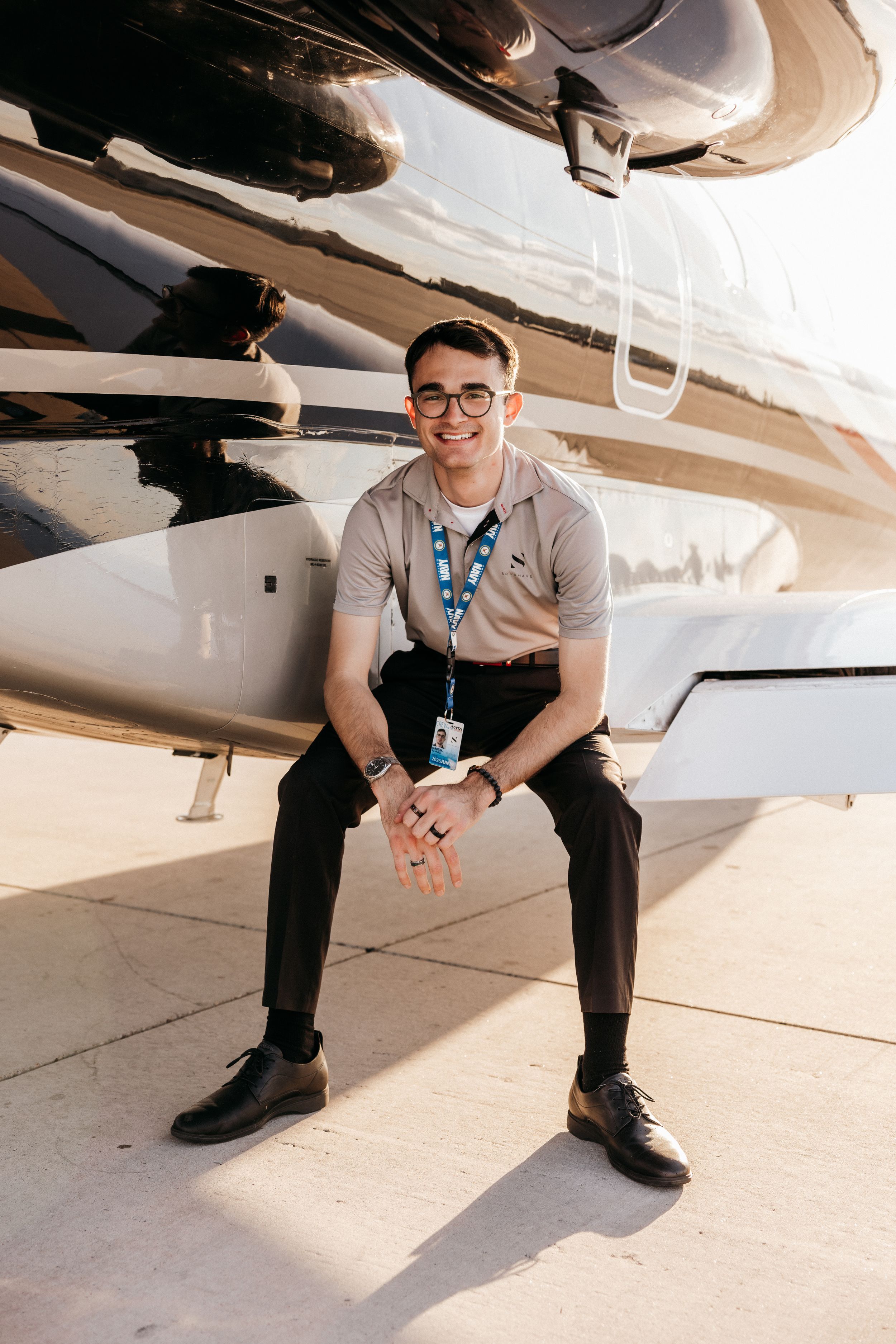 Someone in glasses and a lanyard sits smiling beneath the undercarriage of an aircraft on a sunny day.