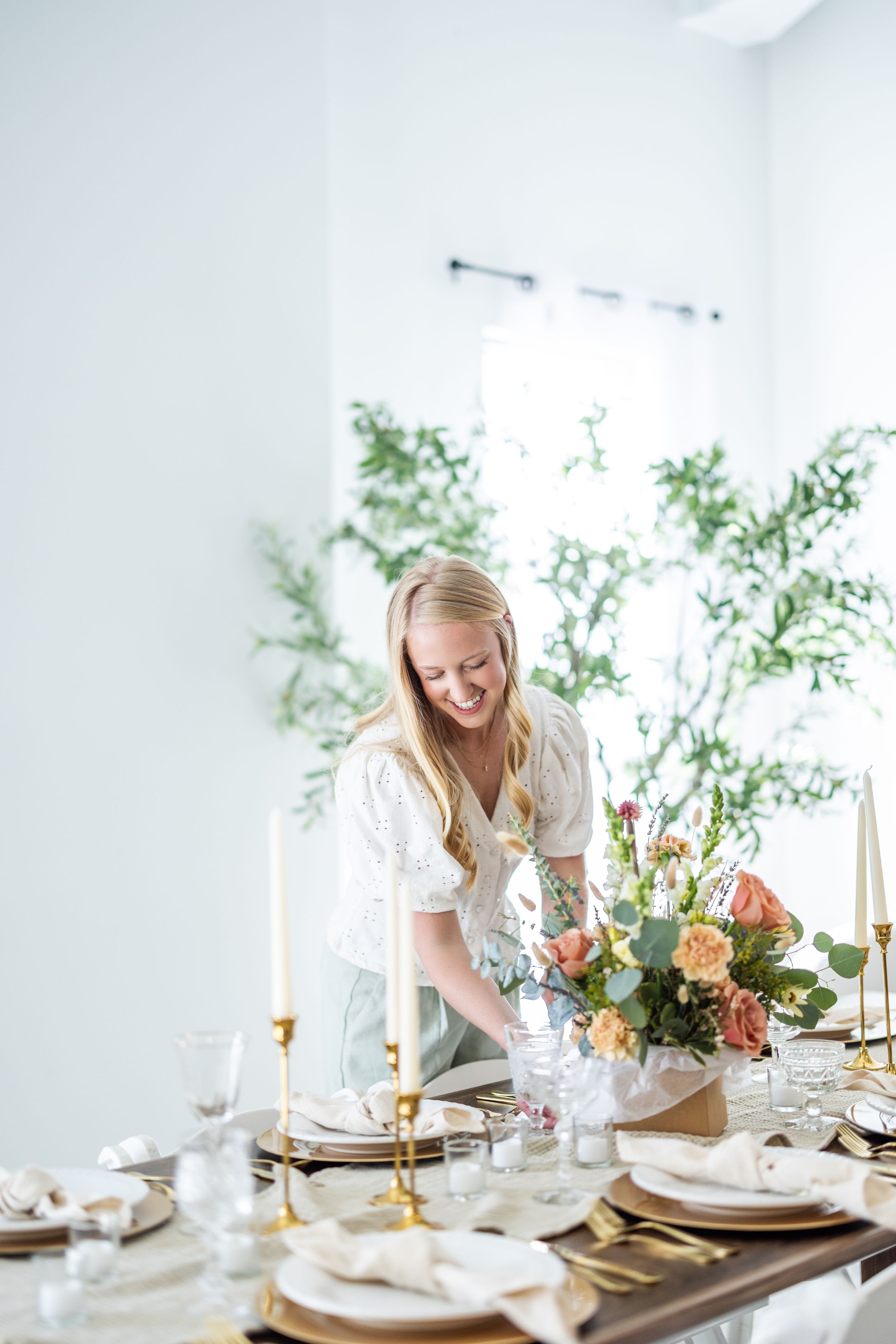 Woman in white arranging elegant table setting with floral centerpiece and gold accents.
