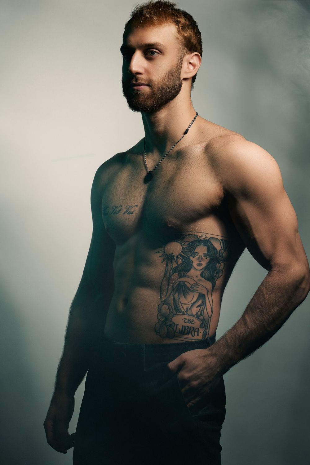 Artistic portrait of muscular shirtless person with beard and large decorative tattoo on torso against moody backdrop.